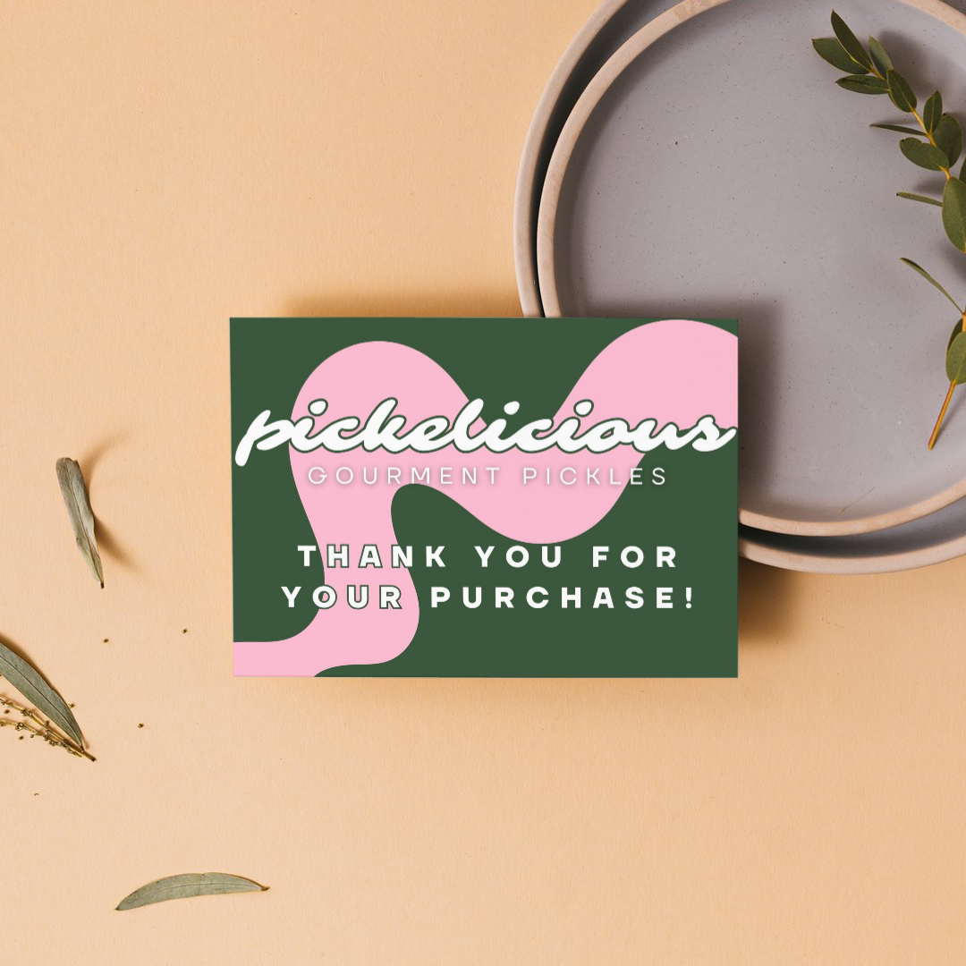 A colorful thank you card on a beige background with ceramic plates and green leaves around it. The card reads ''Picklicious Gourmet Pickles'' with a pink graphic and ''Thank you for your purchase!'' in white uppercase letters.
