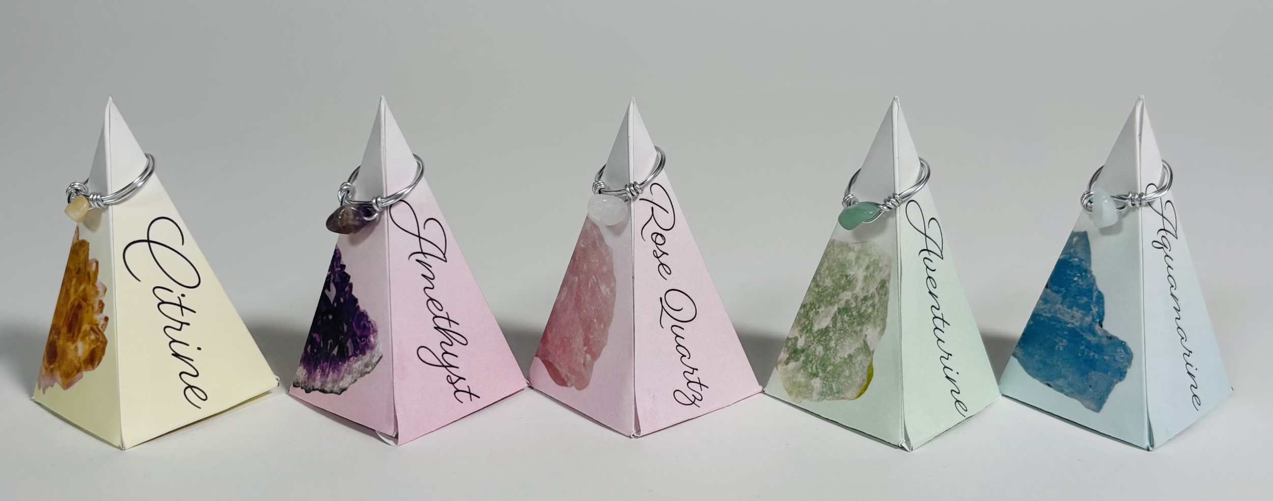 Five pyramid-shaped paper displays with handwritten labels and attached gemstone charms, representing different gemstones: Citrine, Amethyst, Rose Quartz, Green Aventurine, and Aquamarine.