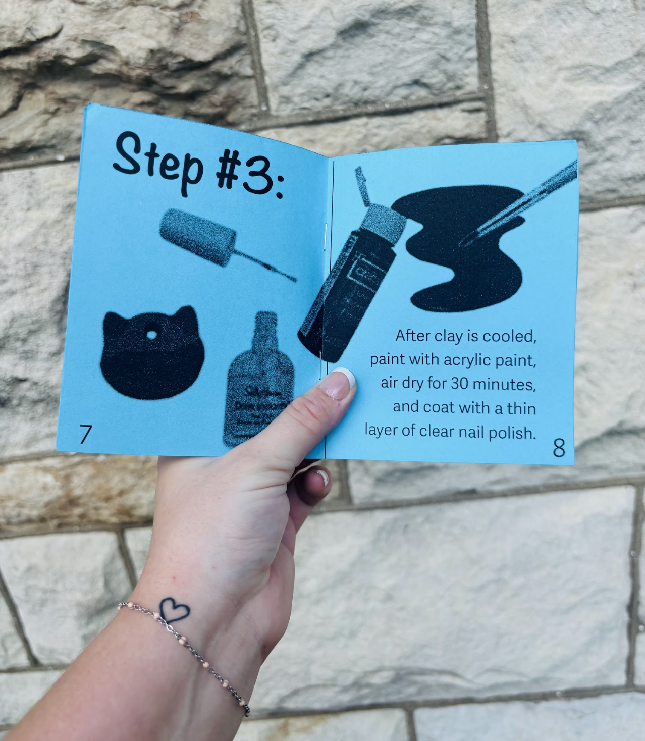 A person holding an open booklet showing step #3 for a craft project. The page includes images of a sponge, nail polish, and paint, along with instructions to paint the clay with acrylic paint after cooling, air dry for 30 minutes, and coat with clea