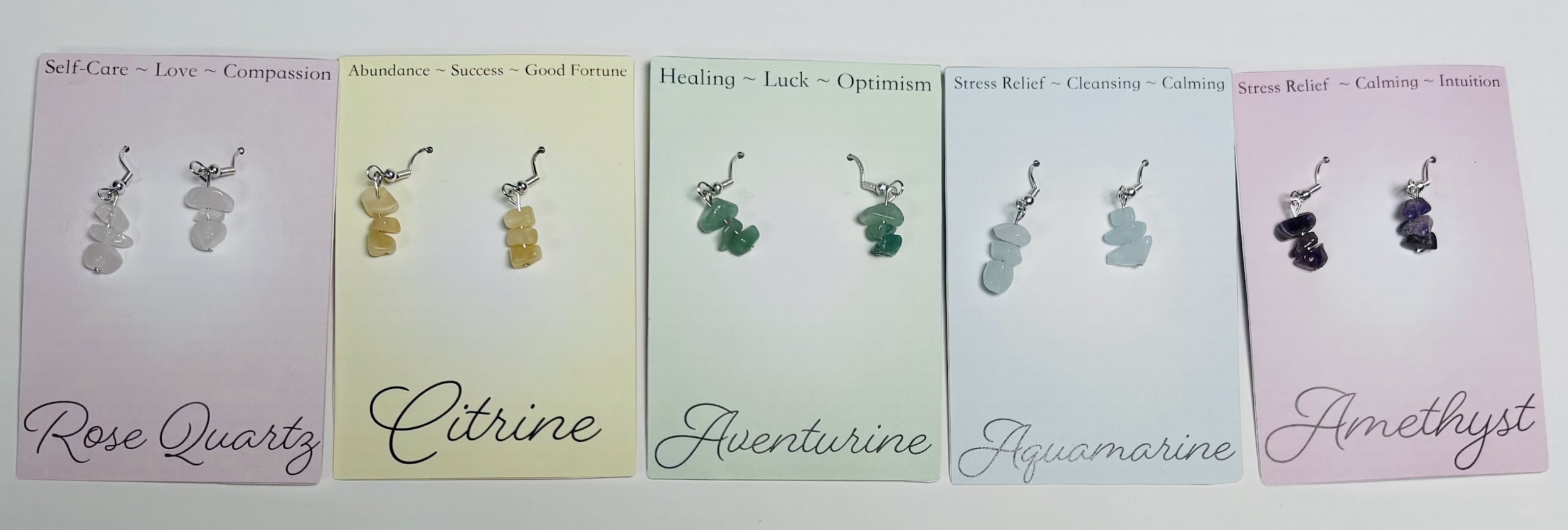 Six pairs of earrings on colored paper cards, each labeled with a gemstone name and associated positive qualities. From left to right: Rose Quartz earrings on pink paper, labeled 'Self-Care, Love, Compassion'; Citrine earrings on yellow paper, labele