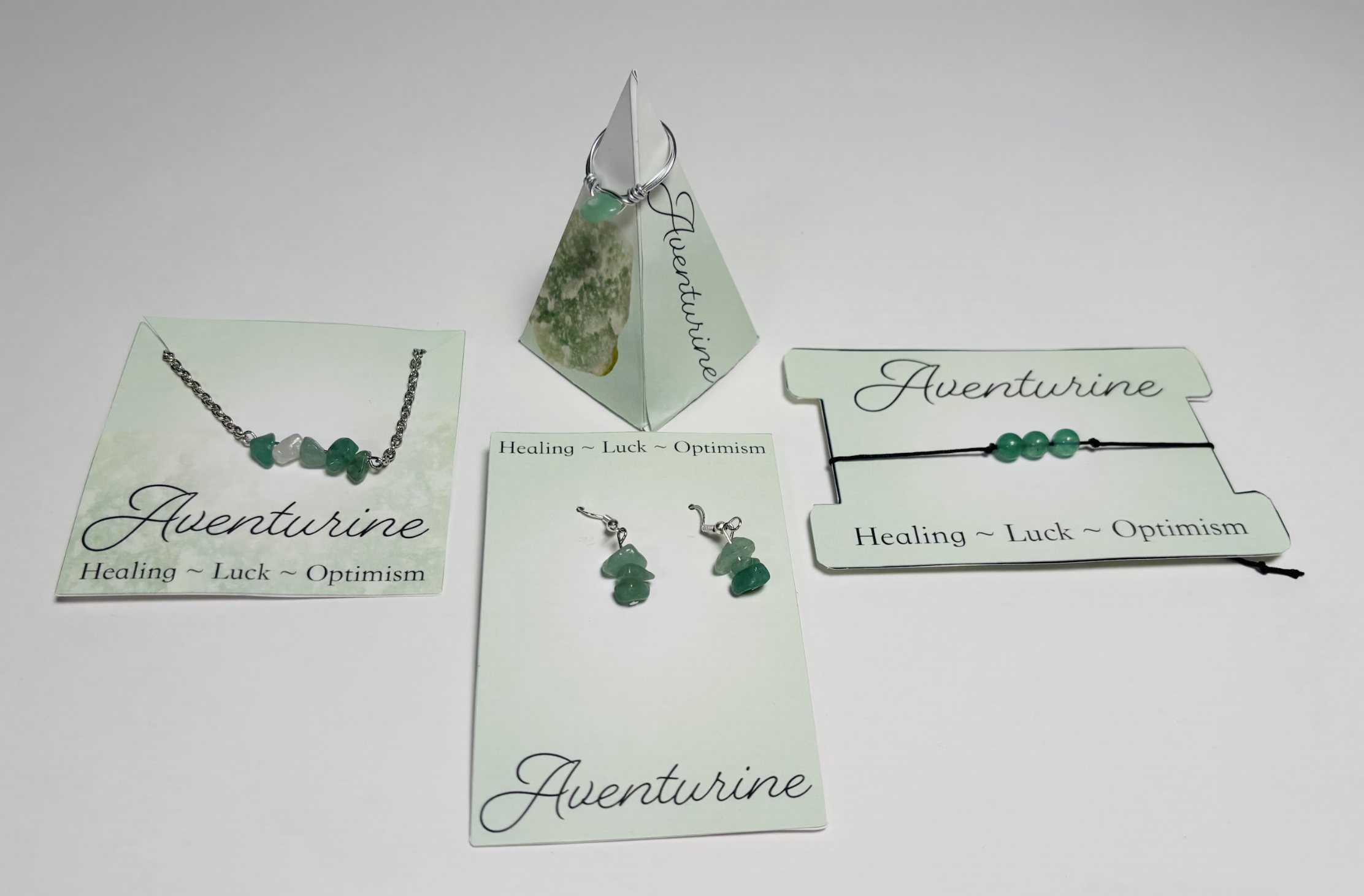 Collection of jewelry items with green stones, including a necklace, earrings, and bracelets, displayed on cards and a pyramid-shaped stand, labeled 'Aventurine' with the words 'Healing ~ Luck ~ Optimism'.