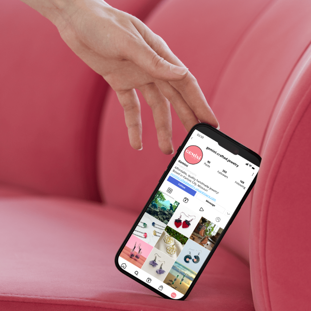 Person's hand reaching towards a smartphone displaying an Instagram profile for 'gemini.crated.jewelry' featuring various colorful jewelry and accessories, with a pink background.