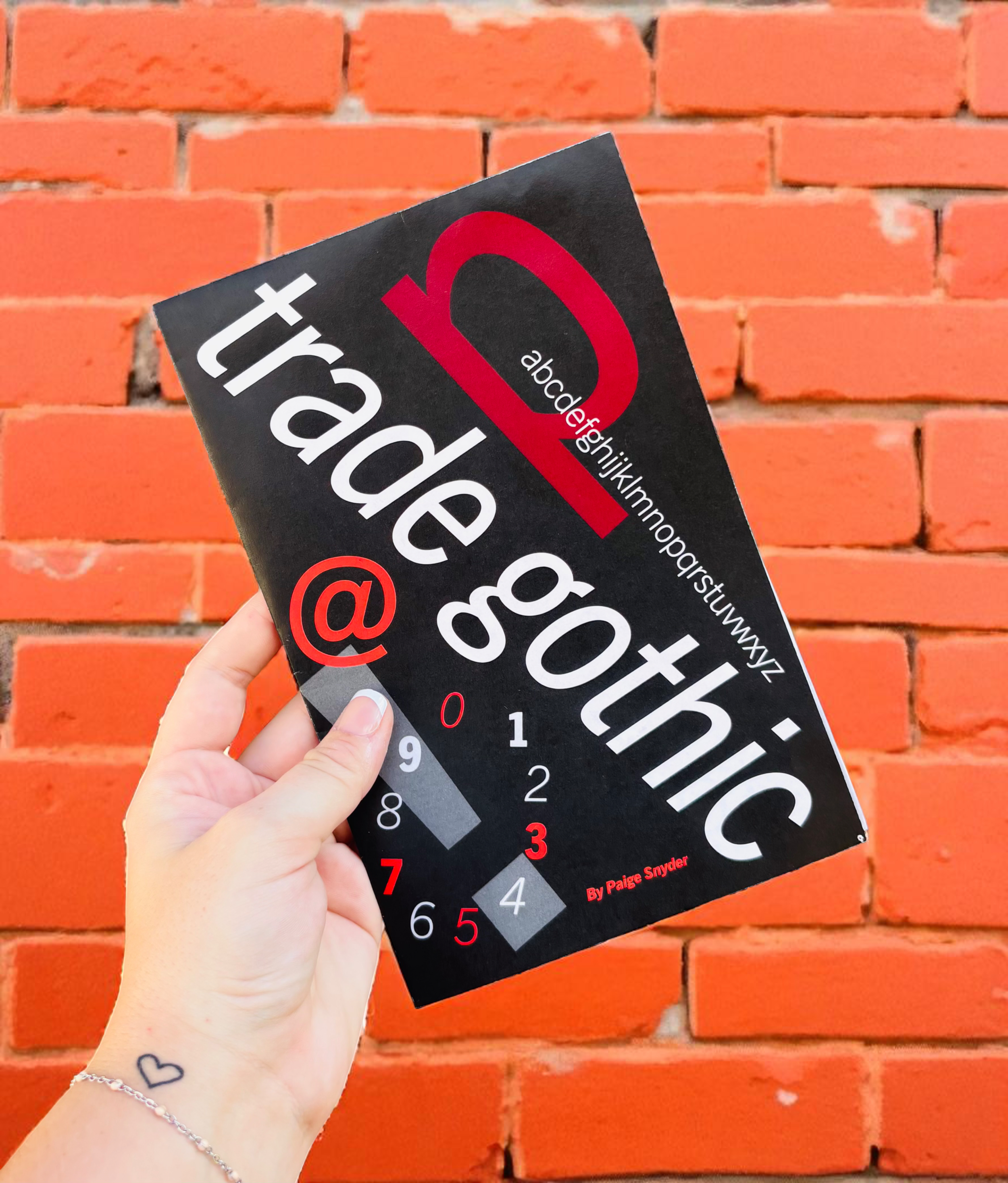 A hand holding a black book titled 'Type Gothic' with red, white, and gray text and design elements, against a red brick wall background.