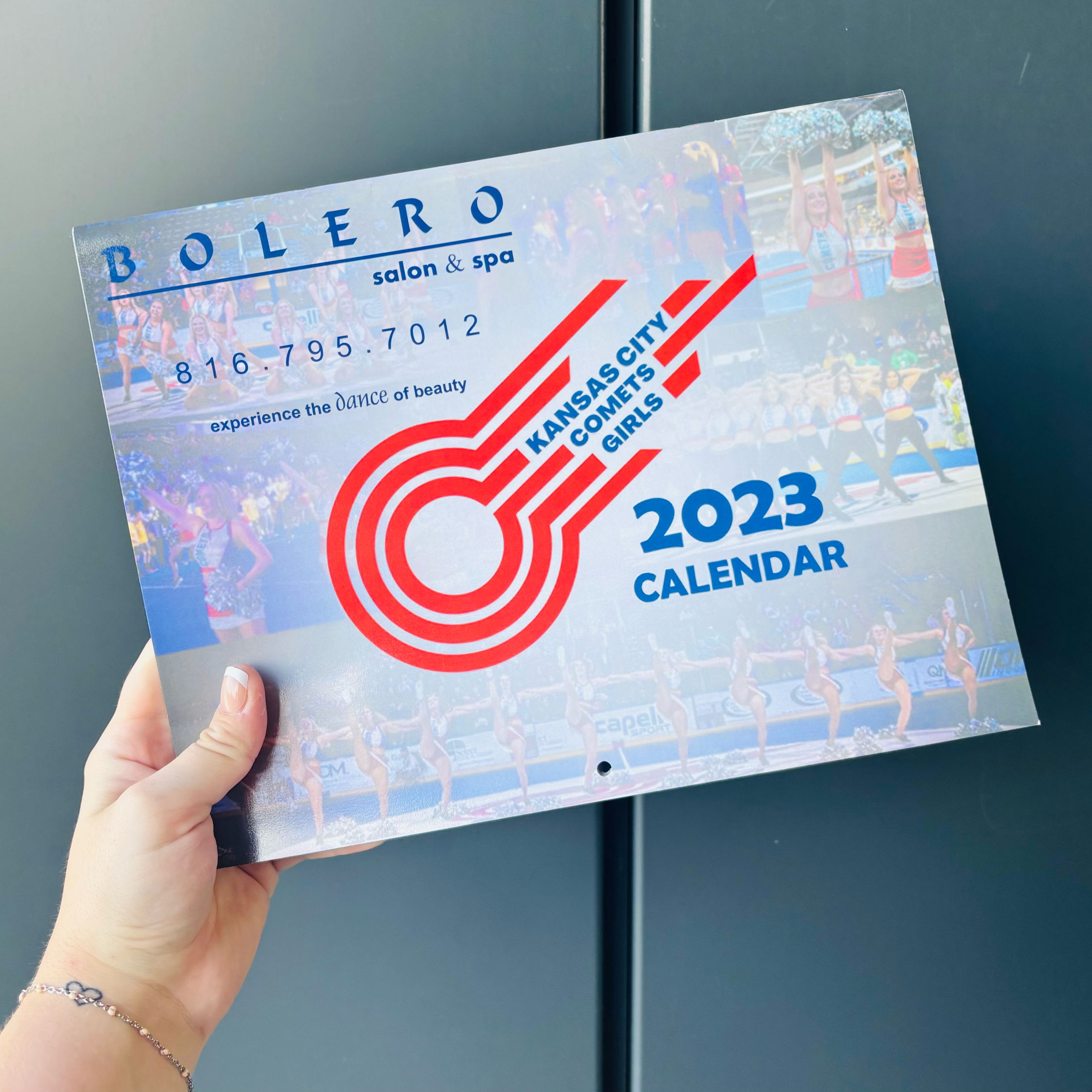 A Kansas City Girls 2023 calendar featuring colorful images of cheerleaders in performance, with a red and blue graphic design and the Kansas City city name, held by a person with a small bracelet on their wrist.