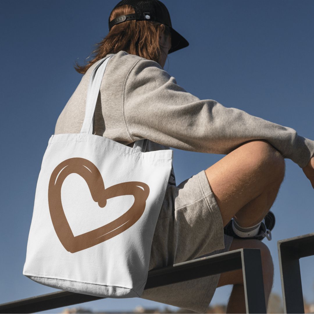 Person sitting outdoors with a tote bag that has a brown heart design, wearing a gray sweatshirt, beige shorts, black cap, and sunglasses, against a clear blue sky.