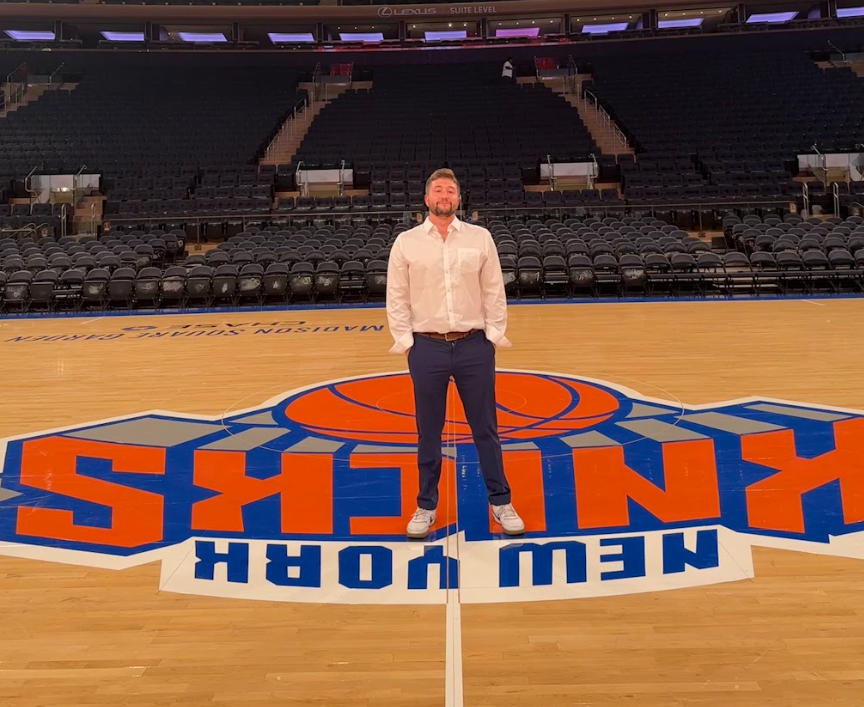 A man standing on the basketball court of Madison Square Garden with a large Knicks logo on the floor. The arena seating is empty in the background.