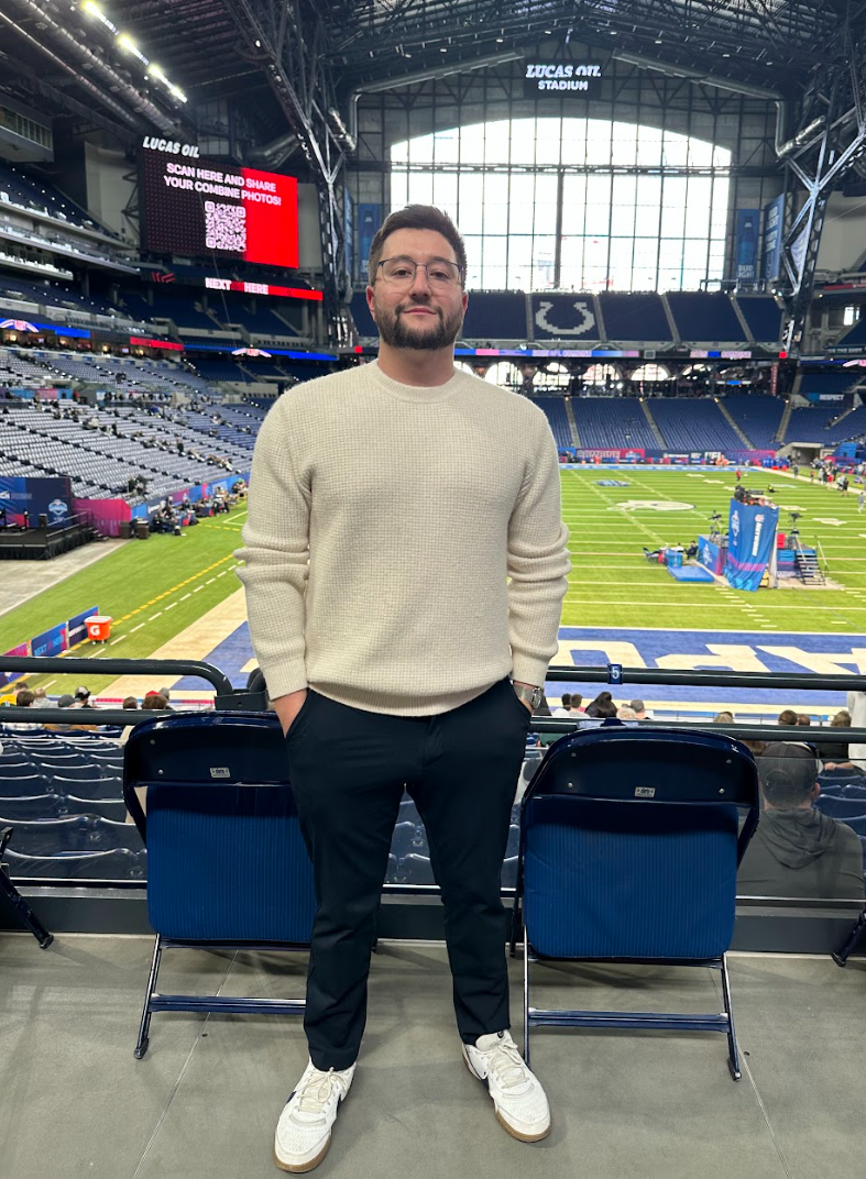A man stands inside a large stadium with a football field visible behind him, featuring empty seats, large windows, and a digital scoreboard.