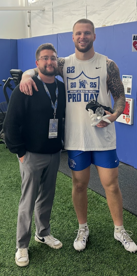 Two men standing inside a sports facility, one wearing a football jersey and shorts, holding gloves, with his arm around the other man in a black sweater and gray pants. Both are smiling.