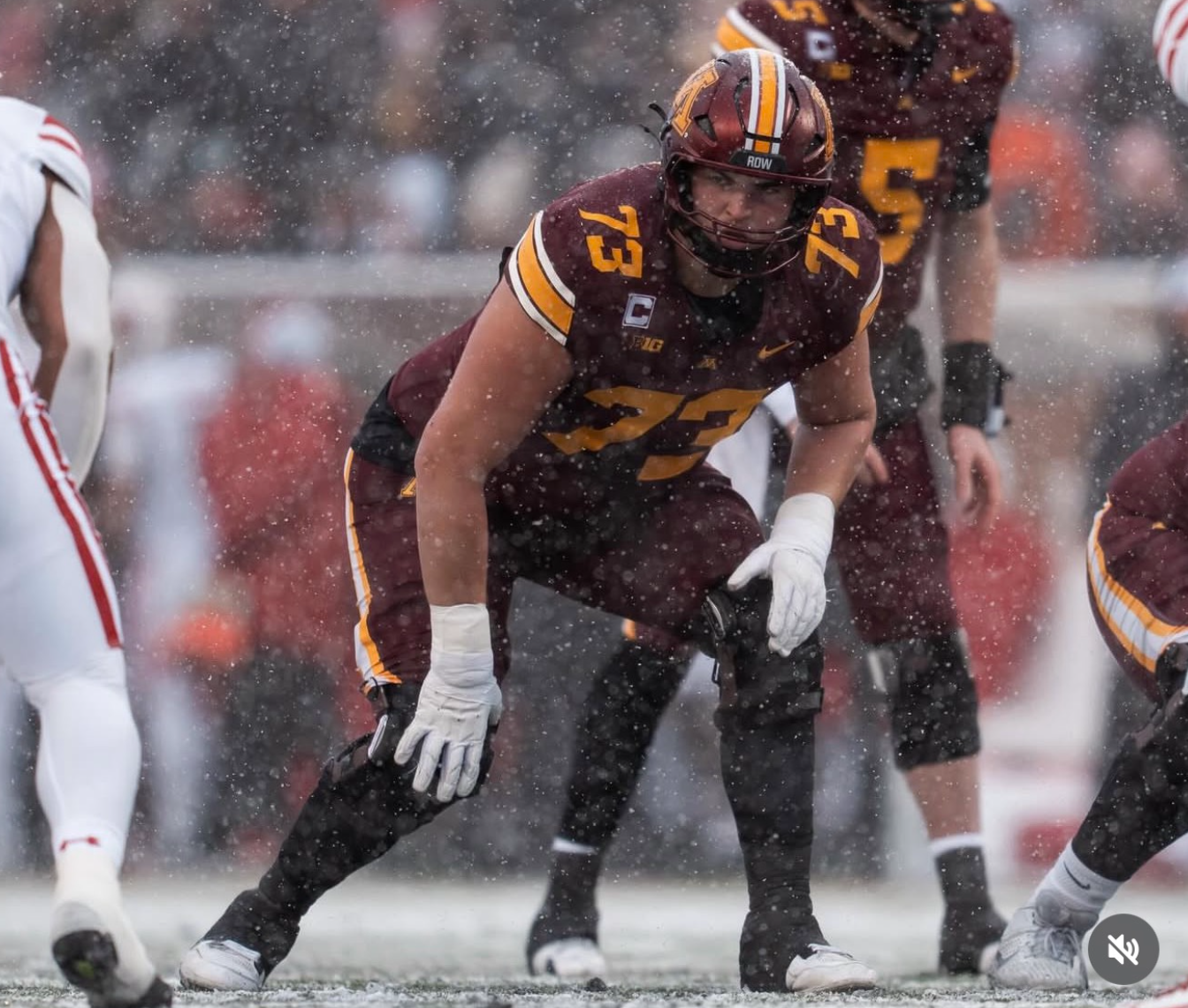 An American football player in a maroon and yellow uniform crouches on the field during a game in the rain, with debris and water droplets visible in the air.