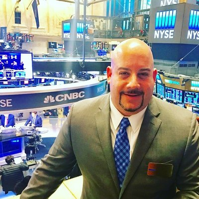 A man in a gray suit and blue tie standing on the trading floor of the New York Stock Exchange (NYSE) with screens and traders in the background.