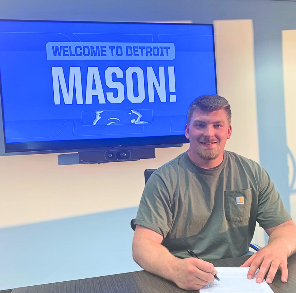 A young man is sitting at a table signing a document, with a large video screen behind him that reads 'Welcome to Detroit Mason!'