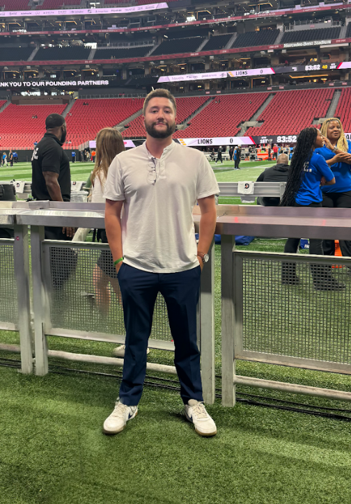 A man standing on the sidelines of a football field inside a stadium, with people in the background and scoreboard showing Lions versus another team.