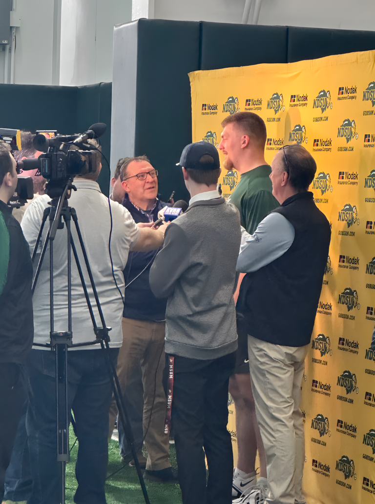 A sports interview taking place in front of a yellow backdrop with the North Dakota State University (NDSU) logo. Several people, including a man in a green shirt and a tall person, are being interviewed by a woman, while a cameraman films the scene.