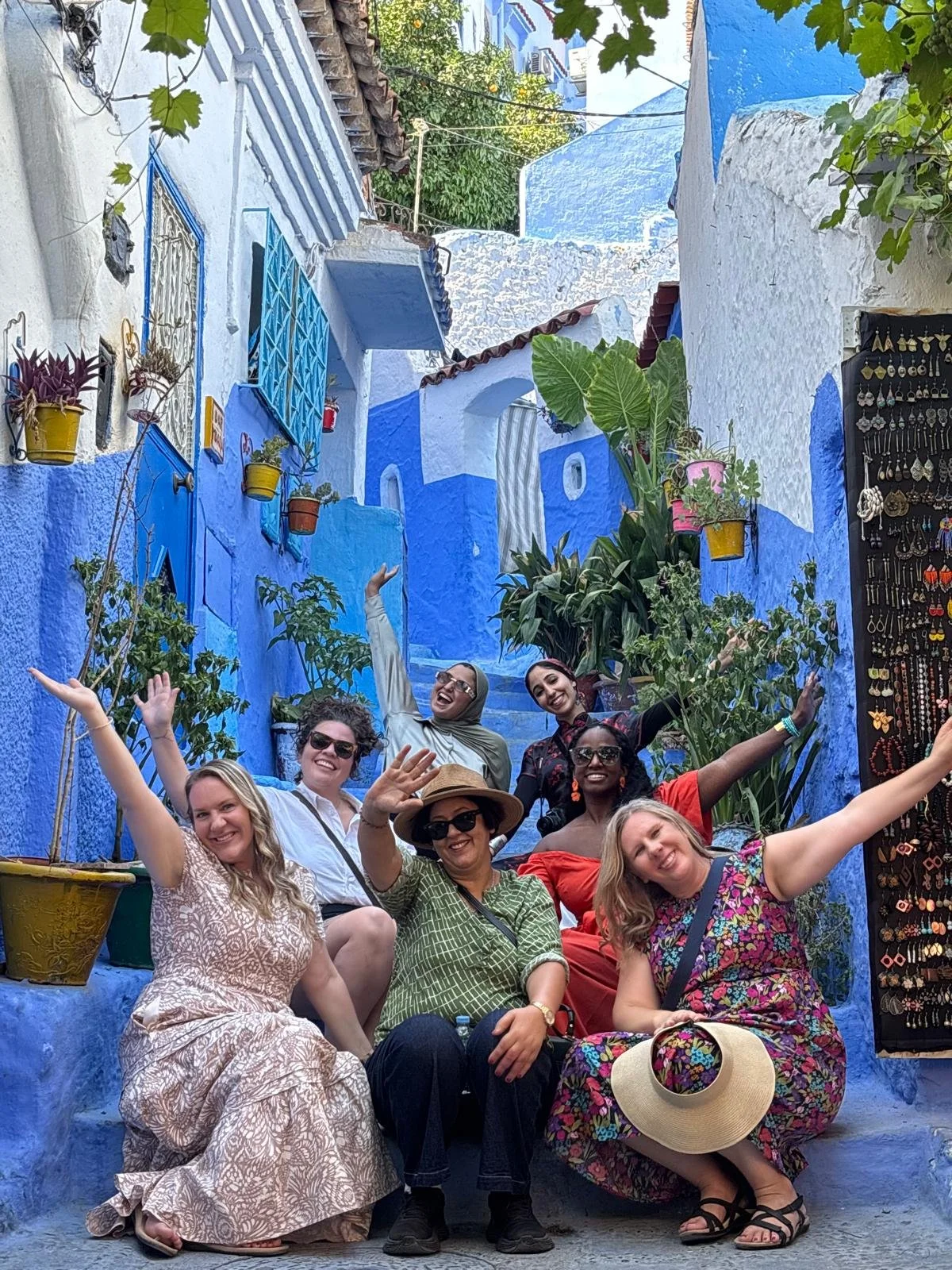 A group of seven women sitting and standing on a colorful blue alleyway with plants, shop, and buildings in the background, all smiling and raising their arms in celebration.