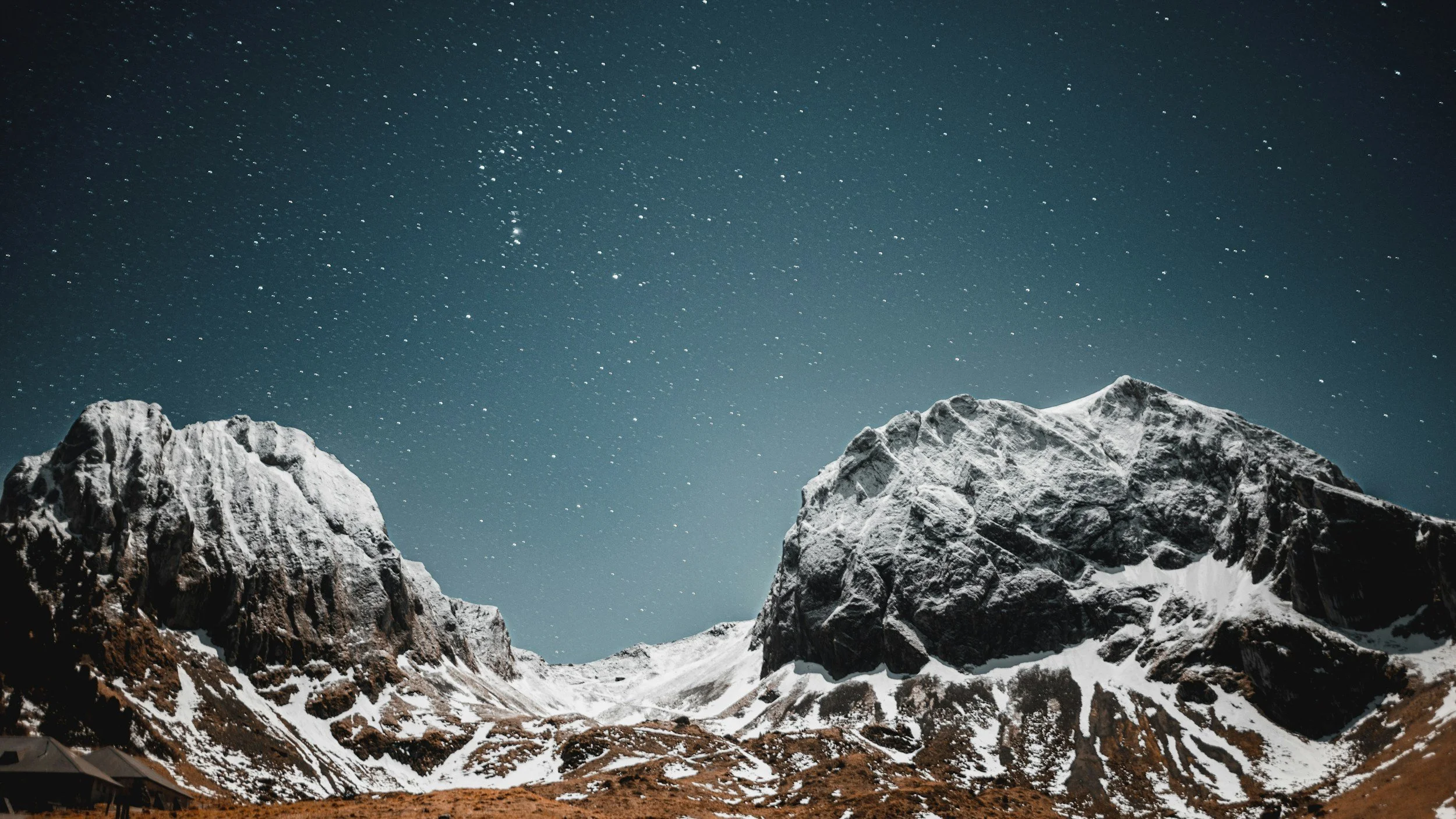 Nighttime mountain scene with snow-covered rugged peaks under a star-filled sky.