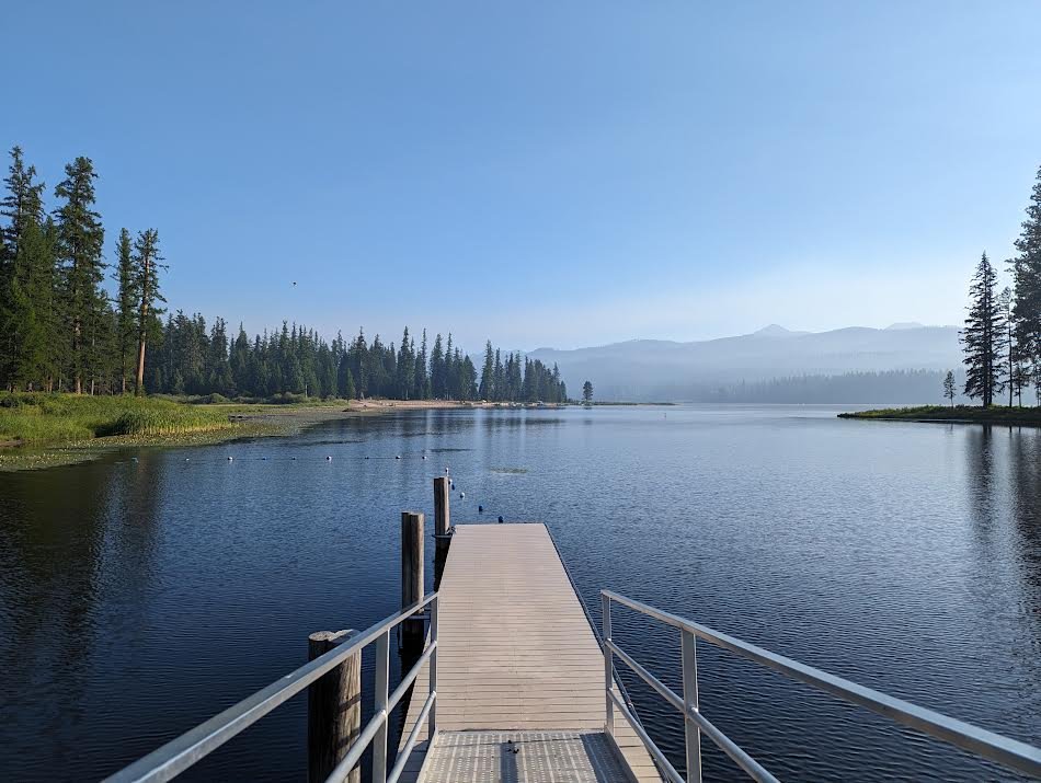 A wooden pier extends into a calm lake surrounded by lush green trees and distant mountains under a clear blue sky.