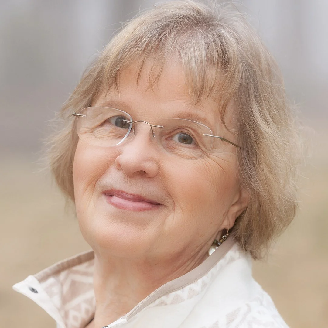 Close-up of an elderly woman with glasses, short blonde hair, and earrings, smiling at the camera.