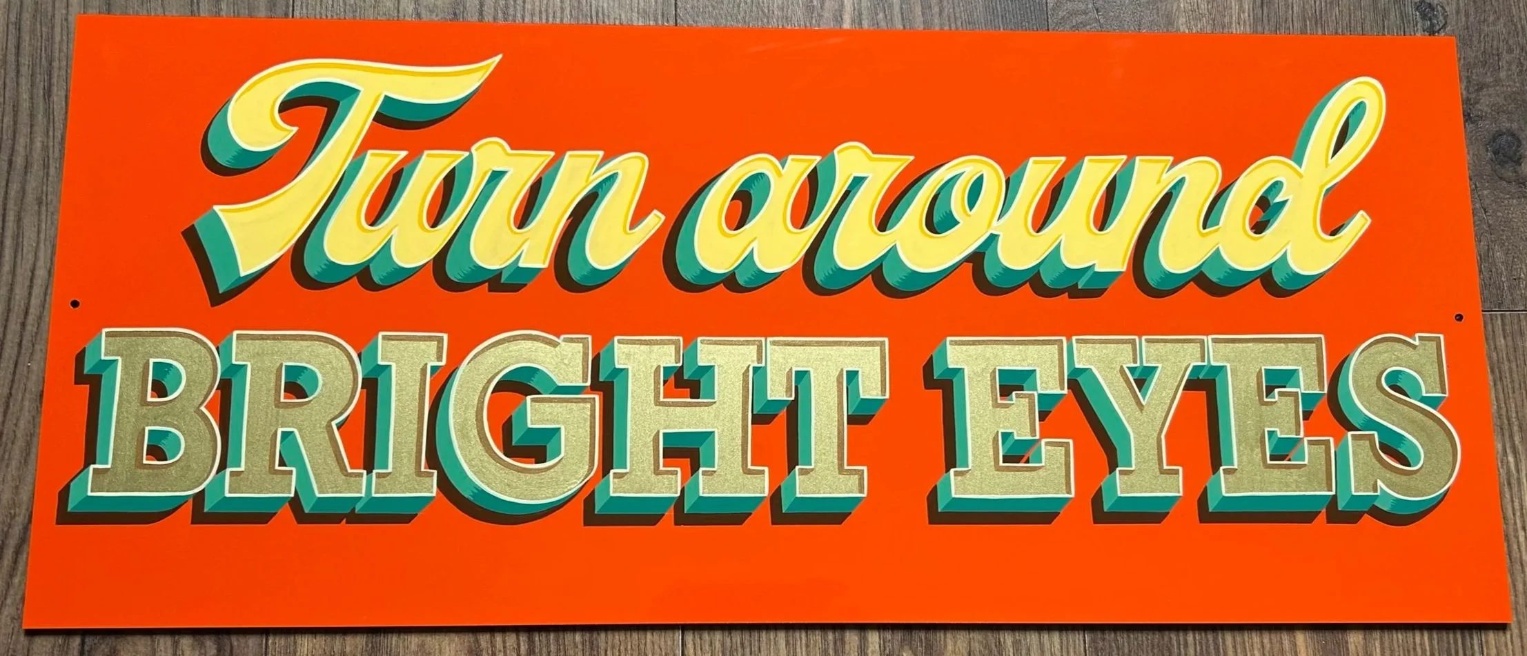 Bright orange sign with colorful 3D letters reading 'Turn around BRIGHT EYES' placed on a wooden floor.