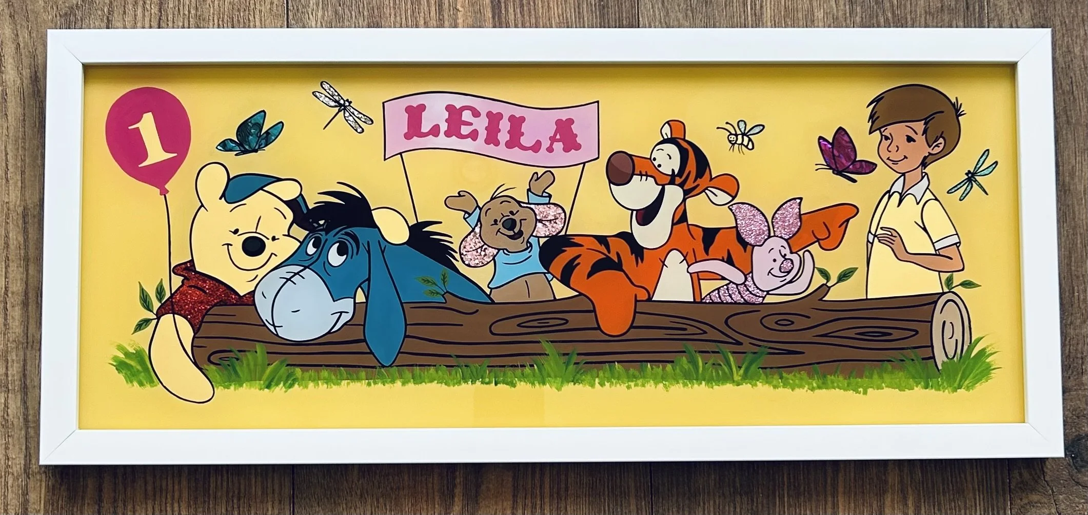 Colorful illustration of Winnie the Pooh, Eeyore, a bear, Tigger, Piglet, and Christopher Robin sitting on a log with a pink banner that says 'Leila' in the center, surrounded by insects and greenery.