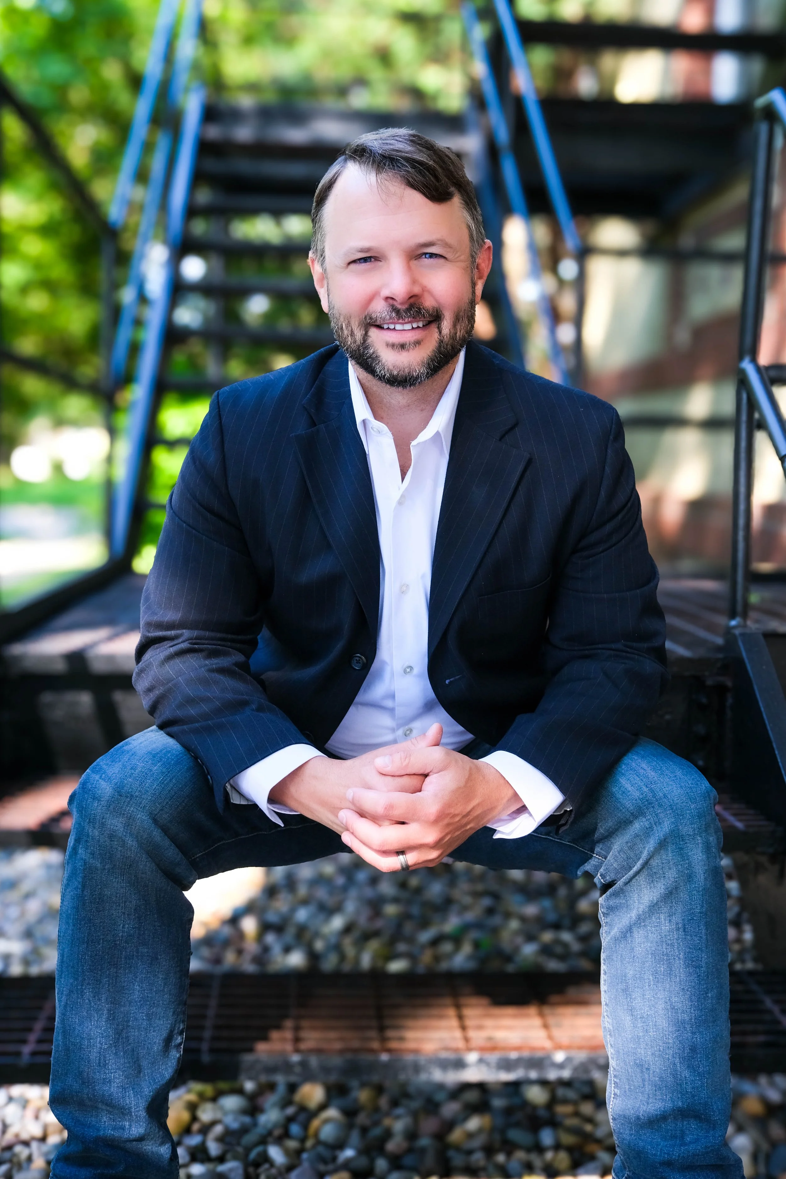 A man in a dark blue blazer and jeans sitting on a metal staircase outdoors, smiling at the camera, with greenery in the background.