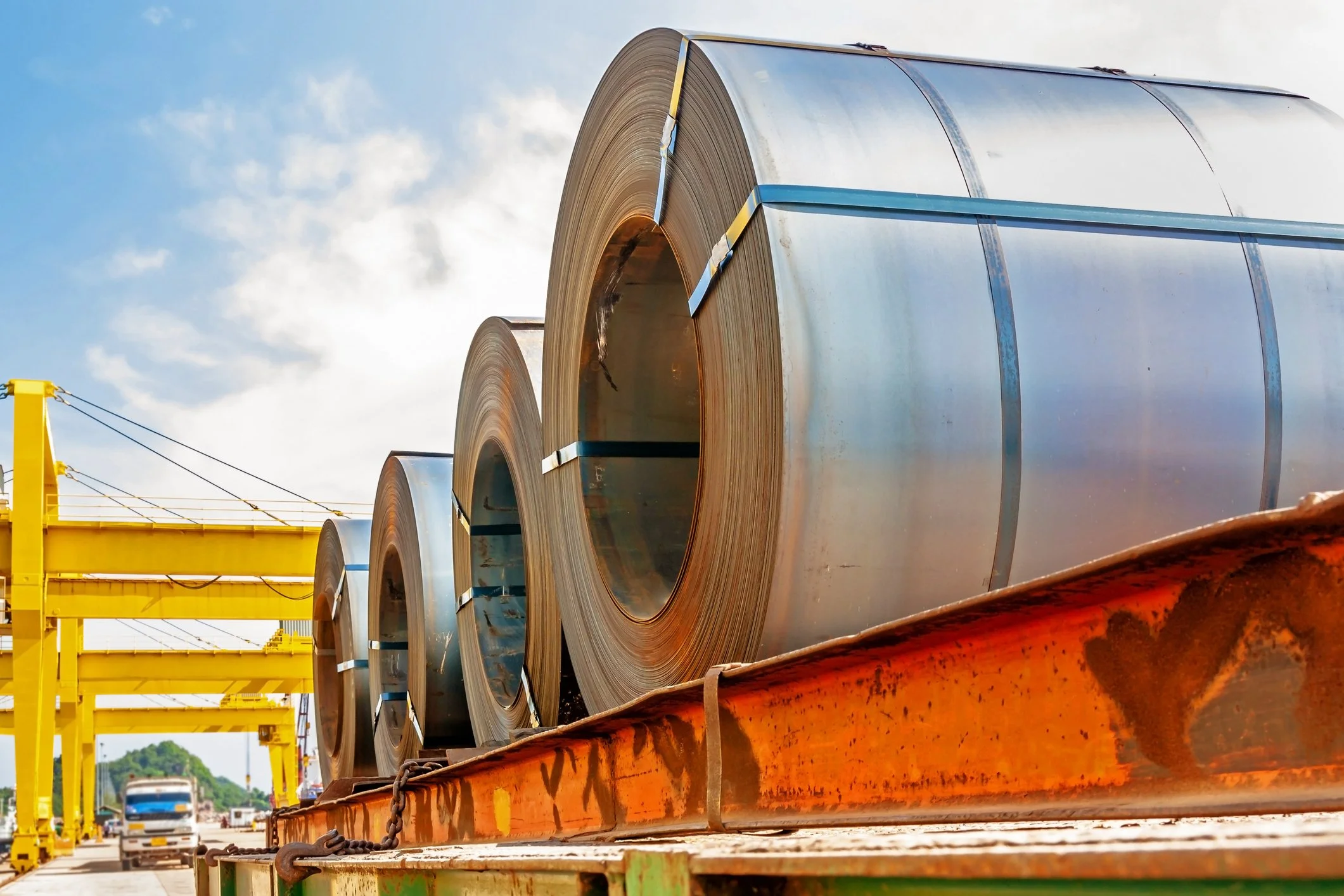 Large steel coils on a flatbed truck at an industrial yard, with yellow cranes and a partly cloudy sky.