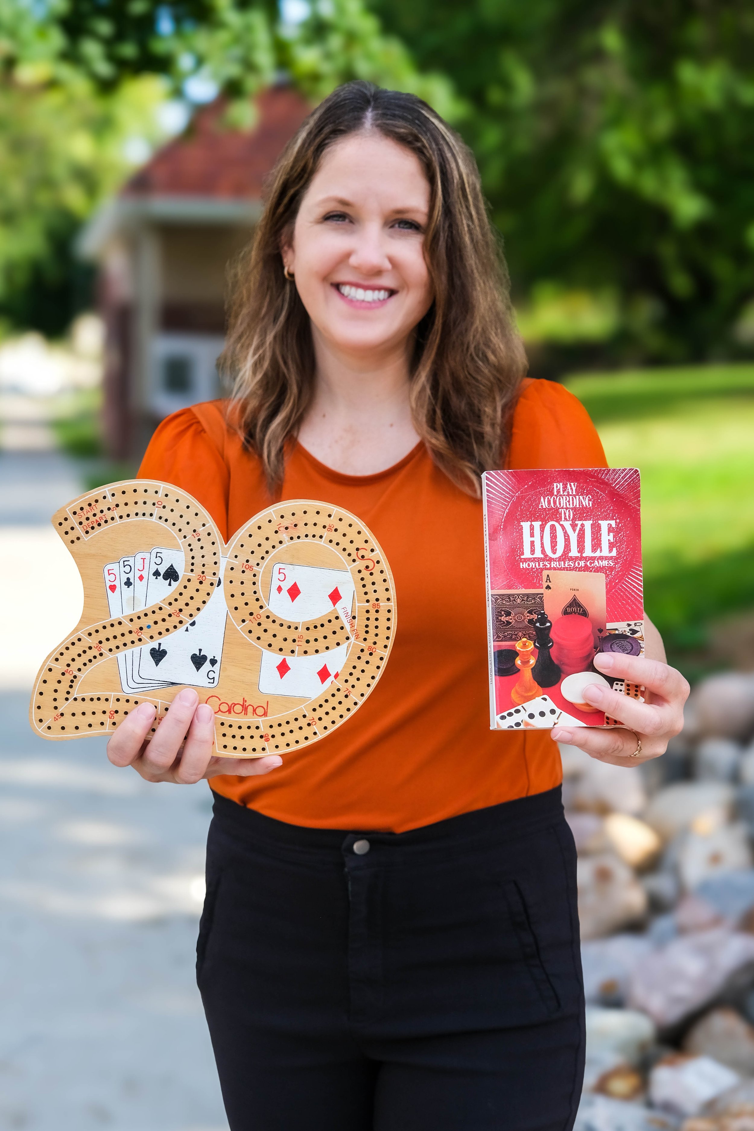 A woman smiling outdoors holding a board game, and a booklet.