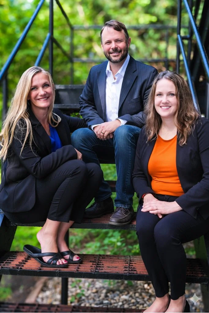 Three people, two women and one man, sitting on a black metal outdoor staircase in a wooded area, smiling at the camera.
