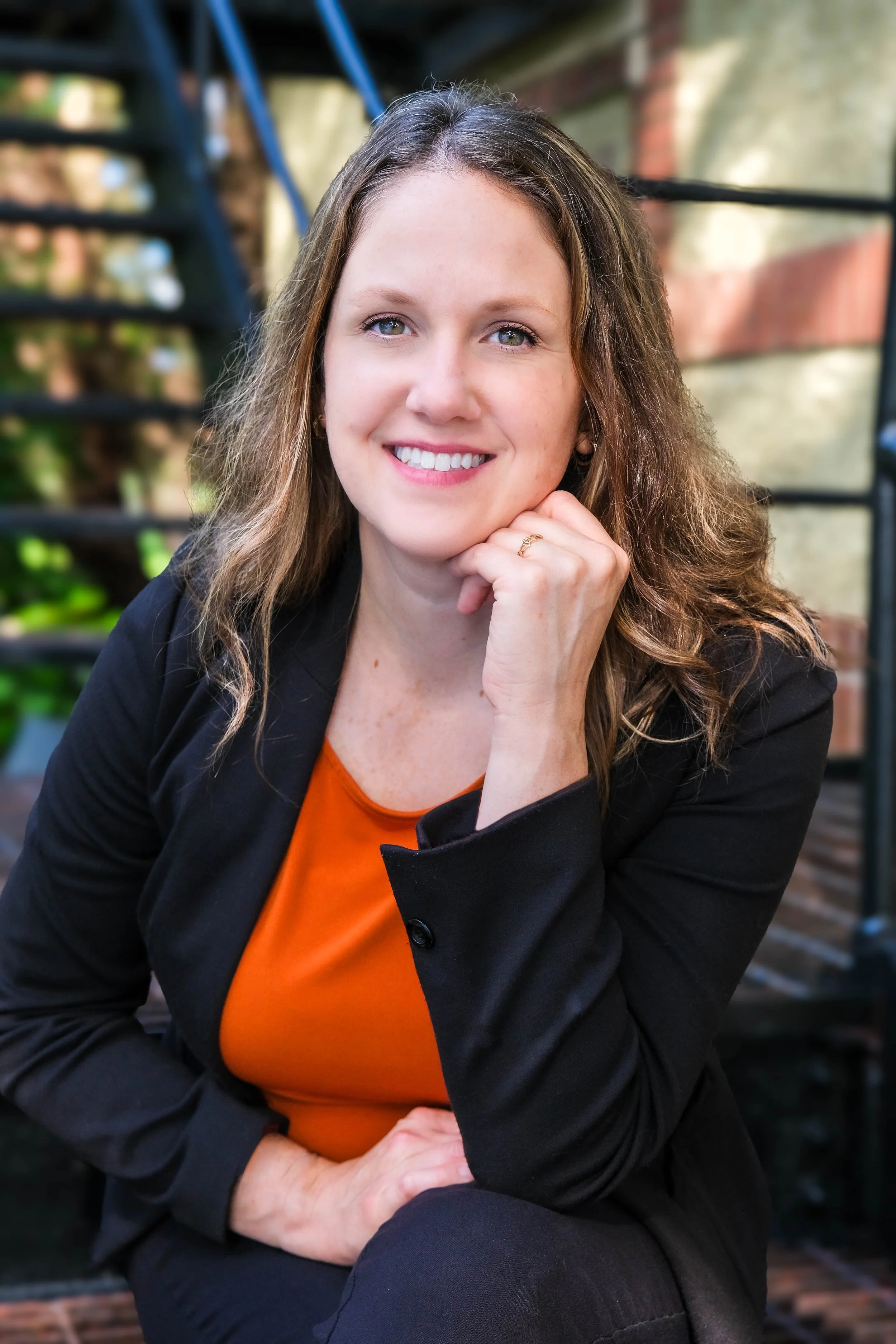 A woman smiling and resting her chin on her hand, wearing a black blazer and an orange top, sitting outdoors.
