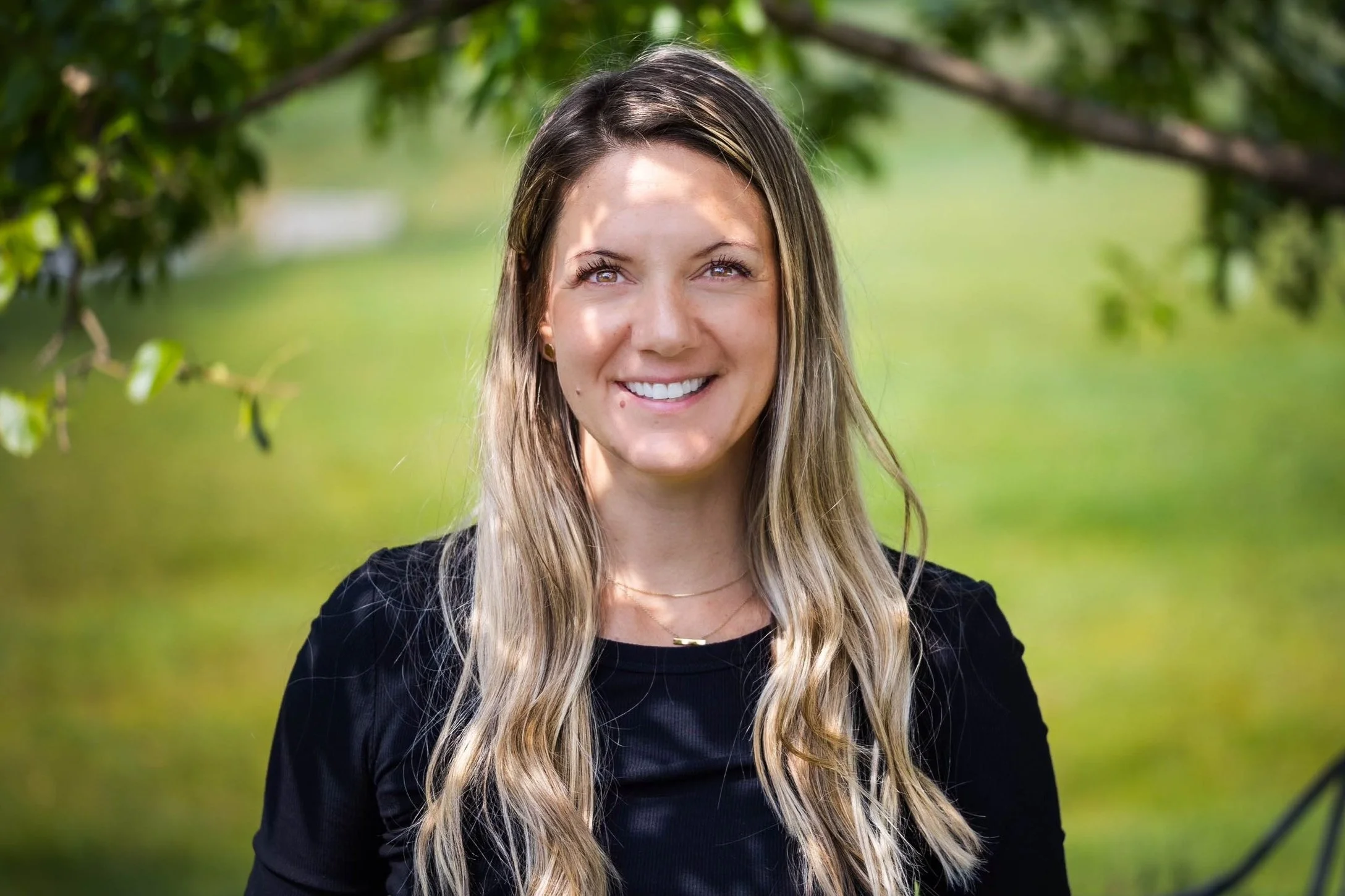 A smiling young woman standing outdoors with green leaves and blurred background.