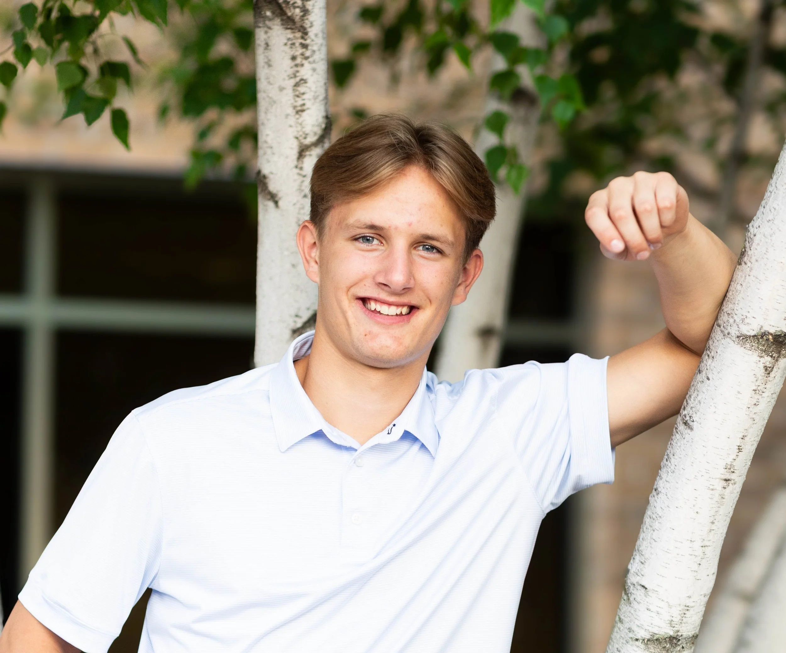 Young man with light brown hair smiling, wearing a white polo shirt, leaning against a white tree branch outdoors.