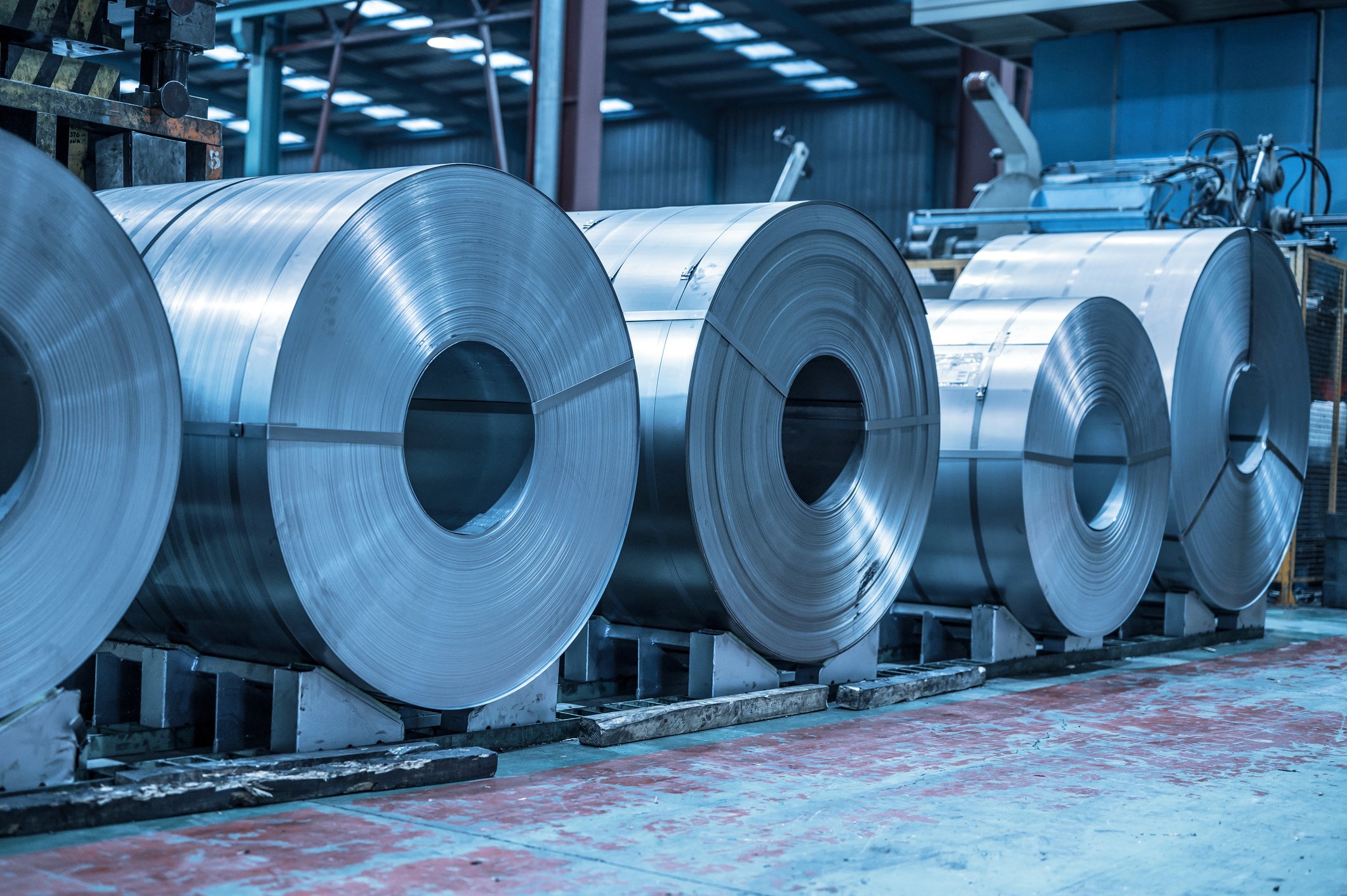 Large rolls of shiny metal sheet stored on pallets in an industrial warehouse.