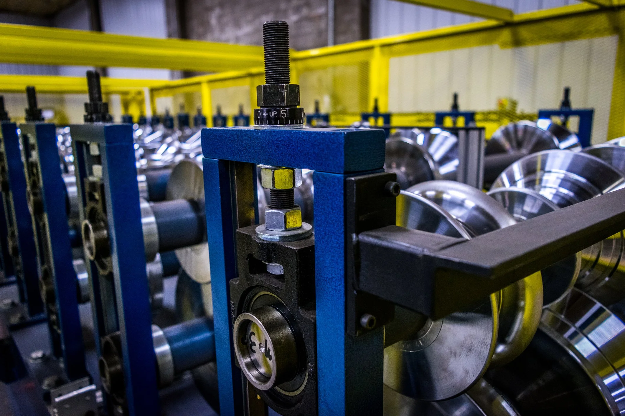 Industrial machinery with metal rollers and a blue frame in a manufacturing setting, with yellow safety barriers overhead.
