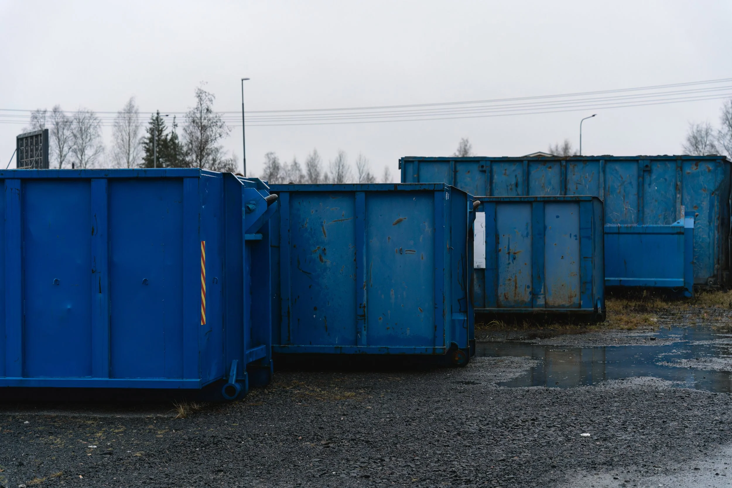 Several large blue industrial dumpsters on a wet, gravelly ground under an overcast sky.