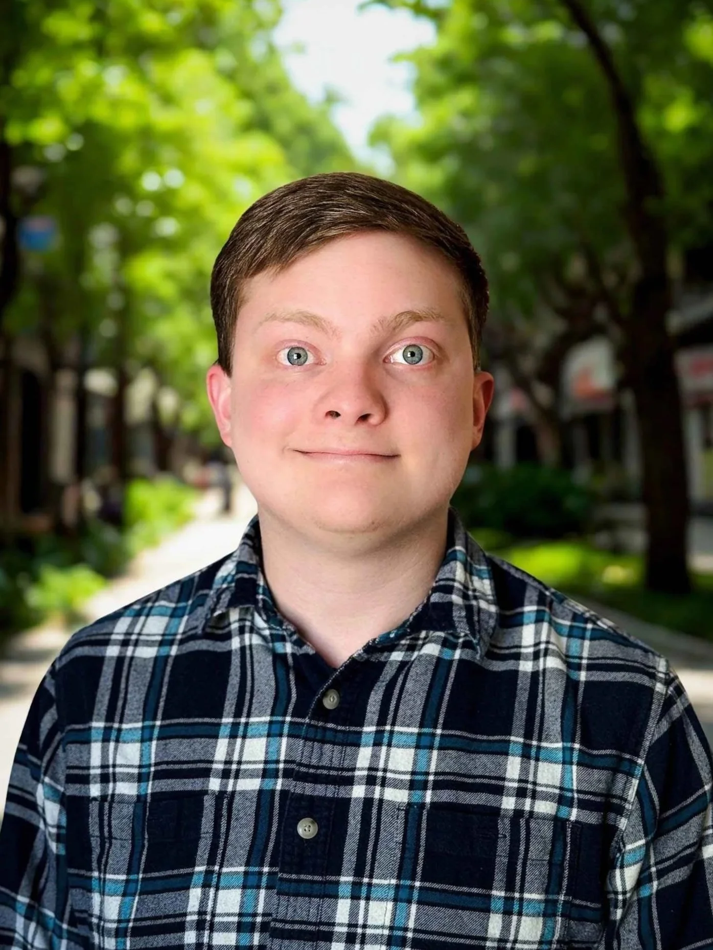 A portrait of a young man with short brown hair, blue eyes, and a playful expression, standing outdoors with green trees and a blurred street background.