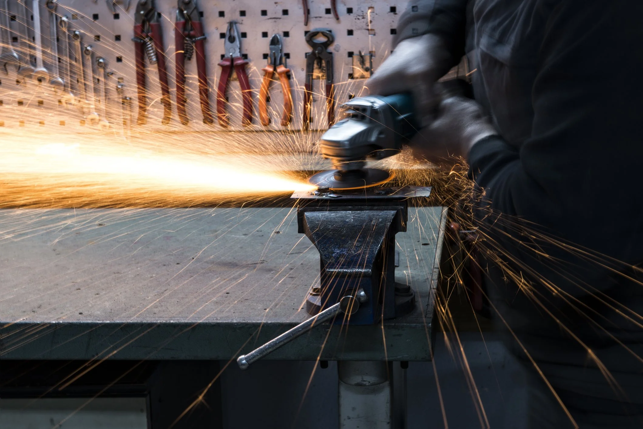 Person grinding metal with an angle grinder, sparks flying, tools hanging on a pegboard in the background.