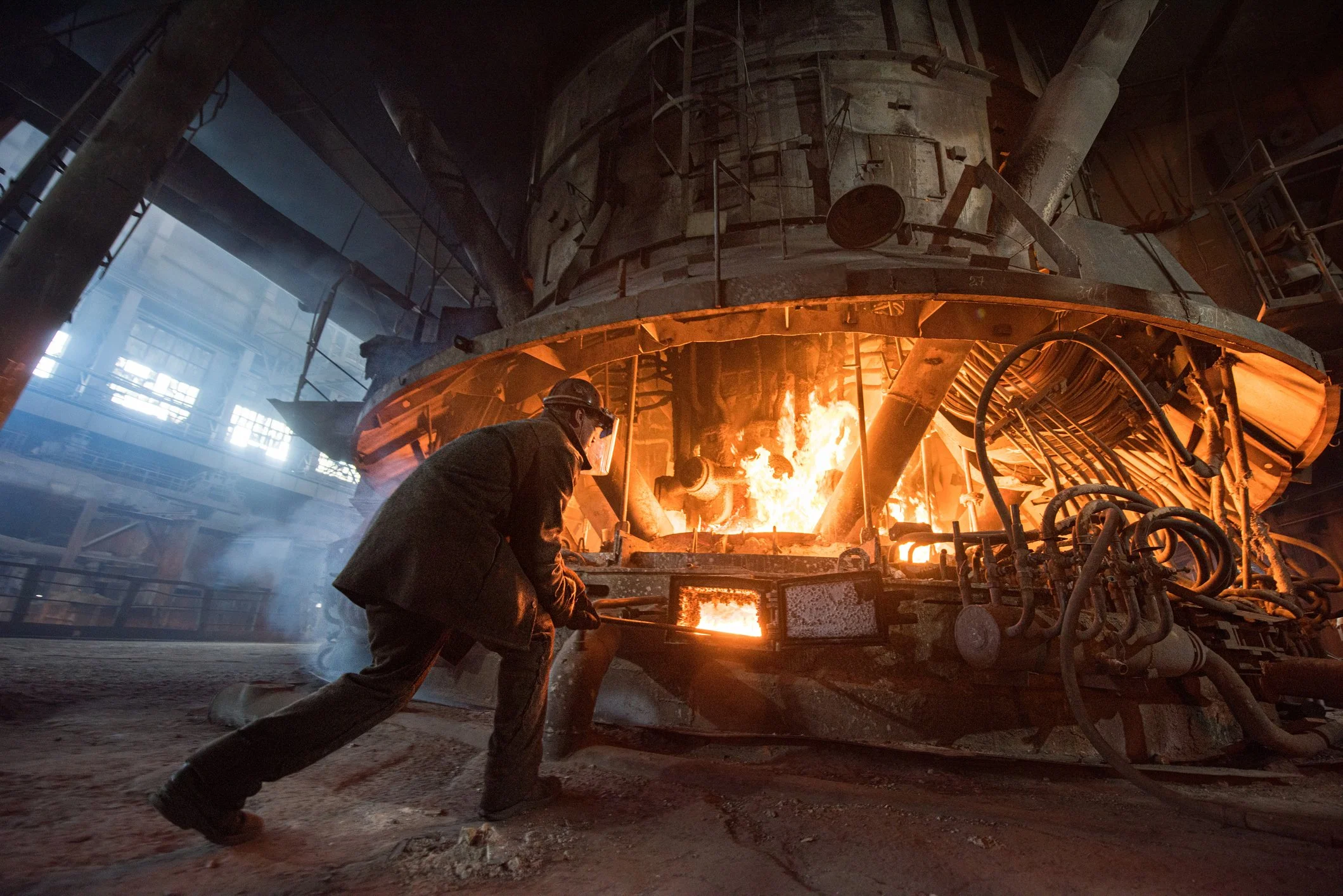 A worker stands near a large industrial furnace that is glowing with fire inside a factory or foundry.