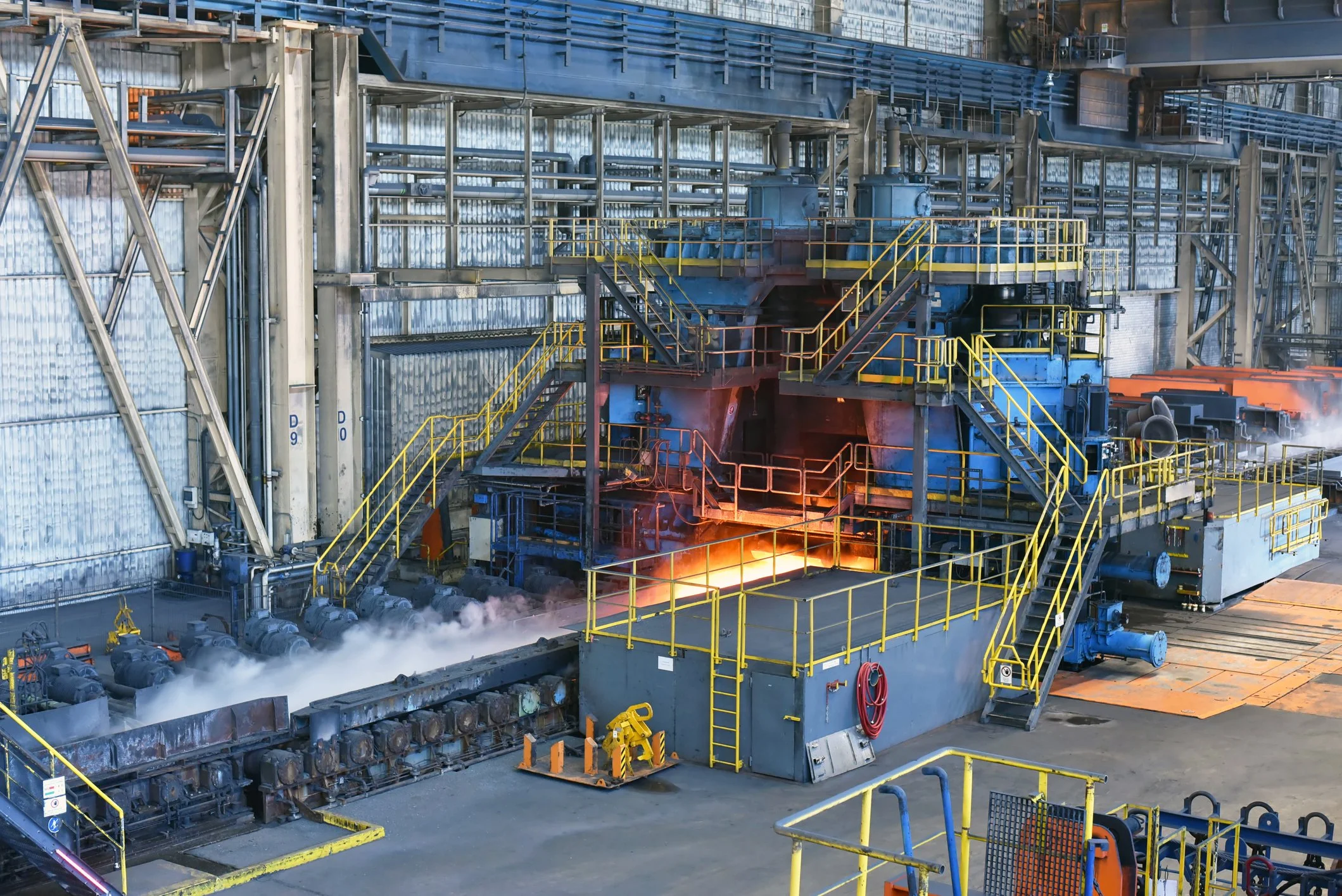 A steel manufacturing plant with molten metal being processed and conveyor belts transporting materials, surrounded by yellow safety railings and industrial equipment.