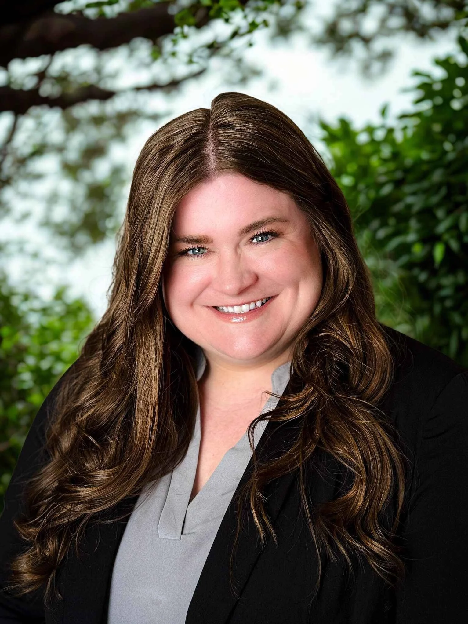 A woman with long wavy brown hair, blue eyes, and a radiant smile, dressed in a light gray top and black blazer, standing outdoors with greenery and trees in the background.