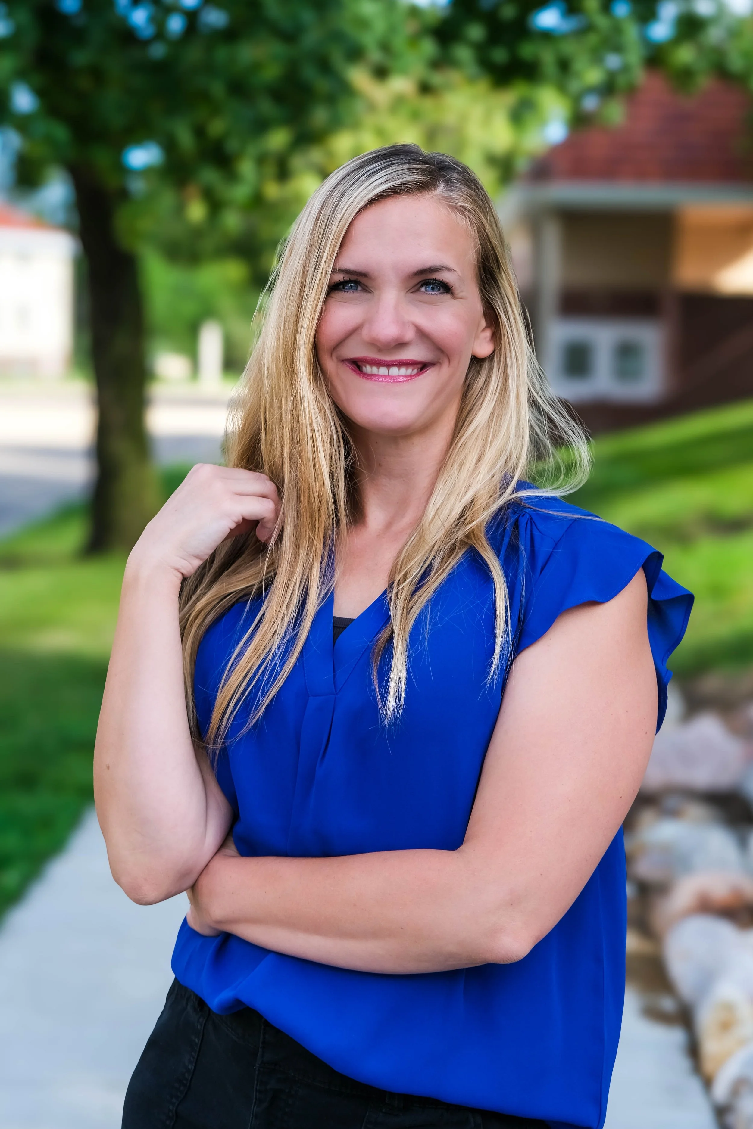 A woman with long blonde hair dressed in a bright blue top standing outdoors on a sidewalk with greenery and trees in the background.