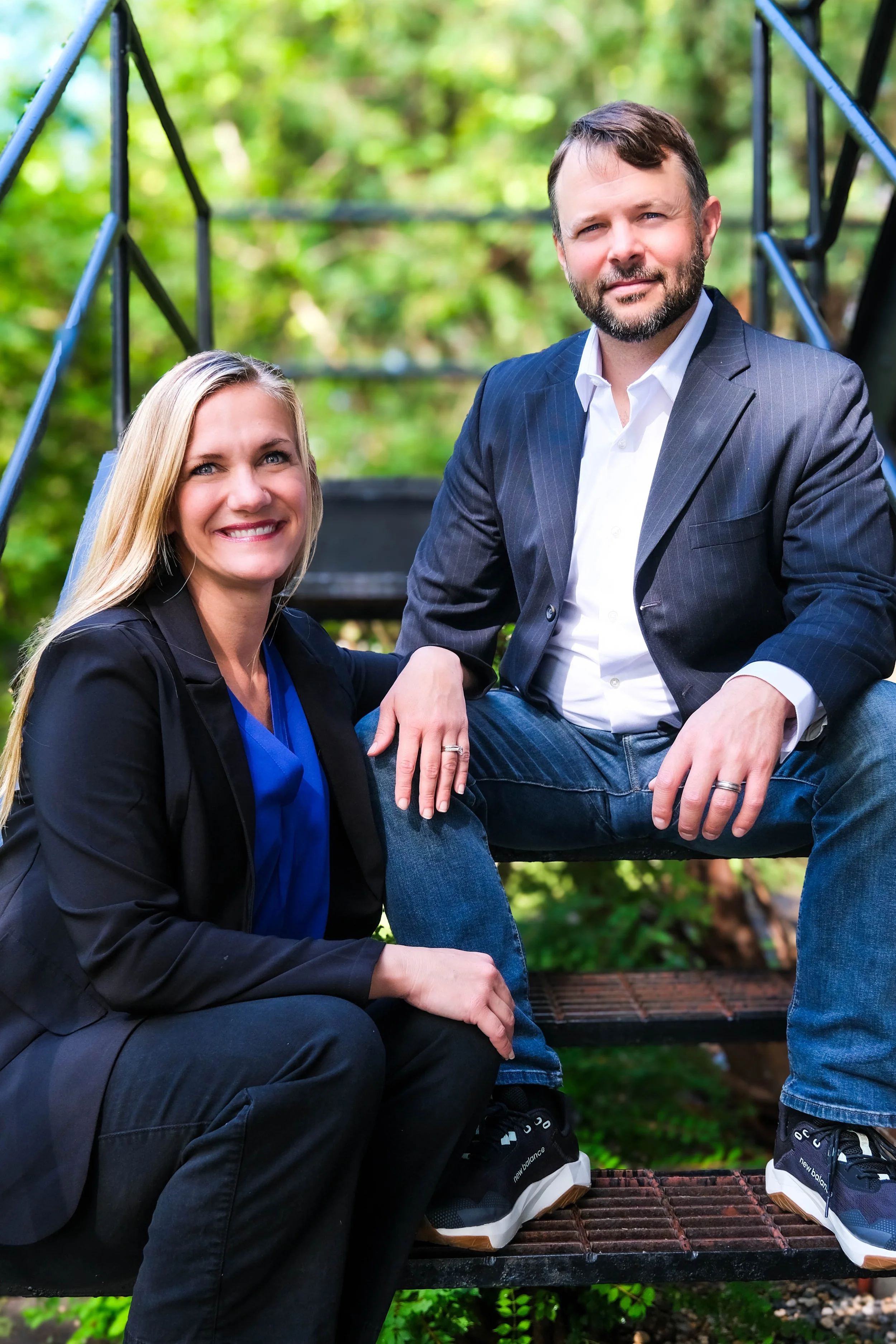 A man and a woman sitting on outdoor metal stairs surrounded by green trees, smiling at the camera.