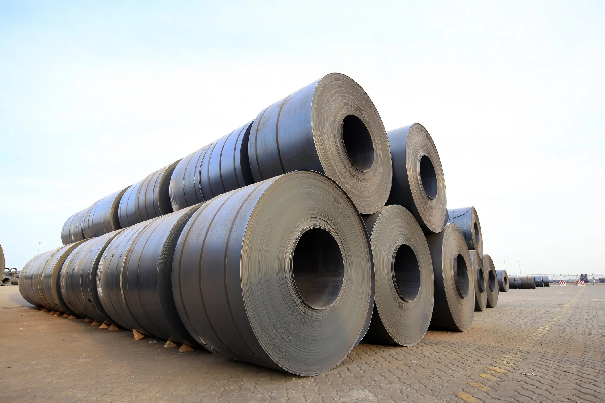 Large rolls of steel coiled on a factory yard with a clear sky in the background.