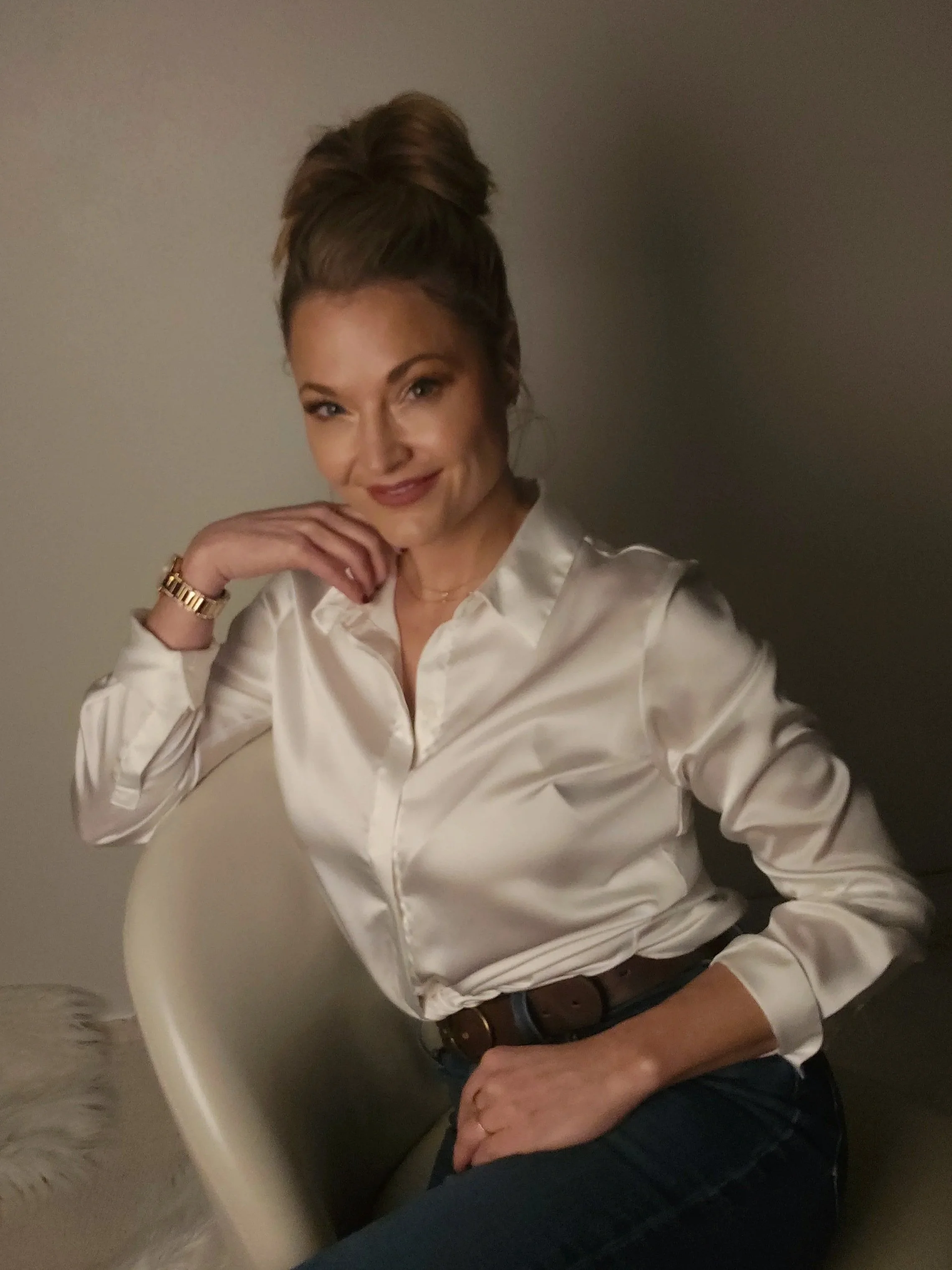 Jasmin Nass sitting on a chair, smiling with her hand near her face, representing calm confidence, clarity, and trust in guided hypnosis.
