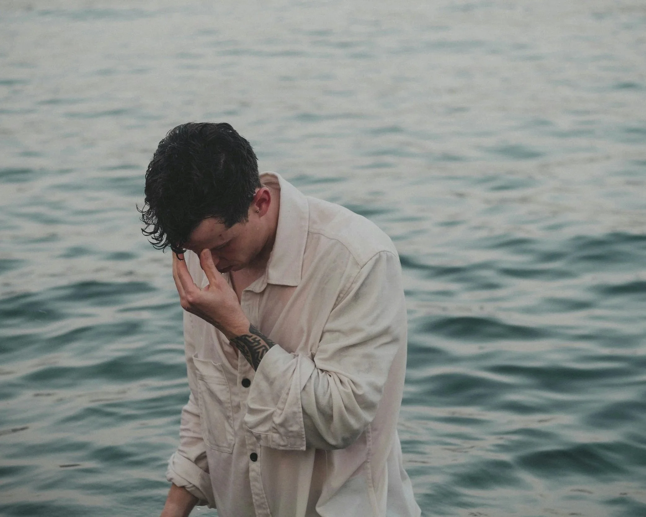 A man stands near the water with his hand on his face, reflecting a moment of emotional overwhelm.