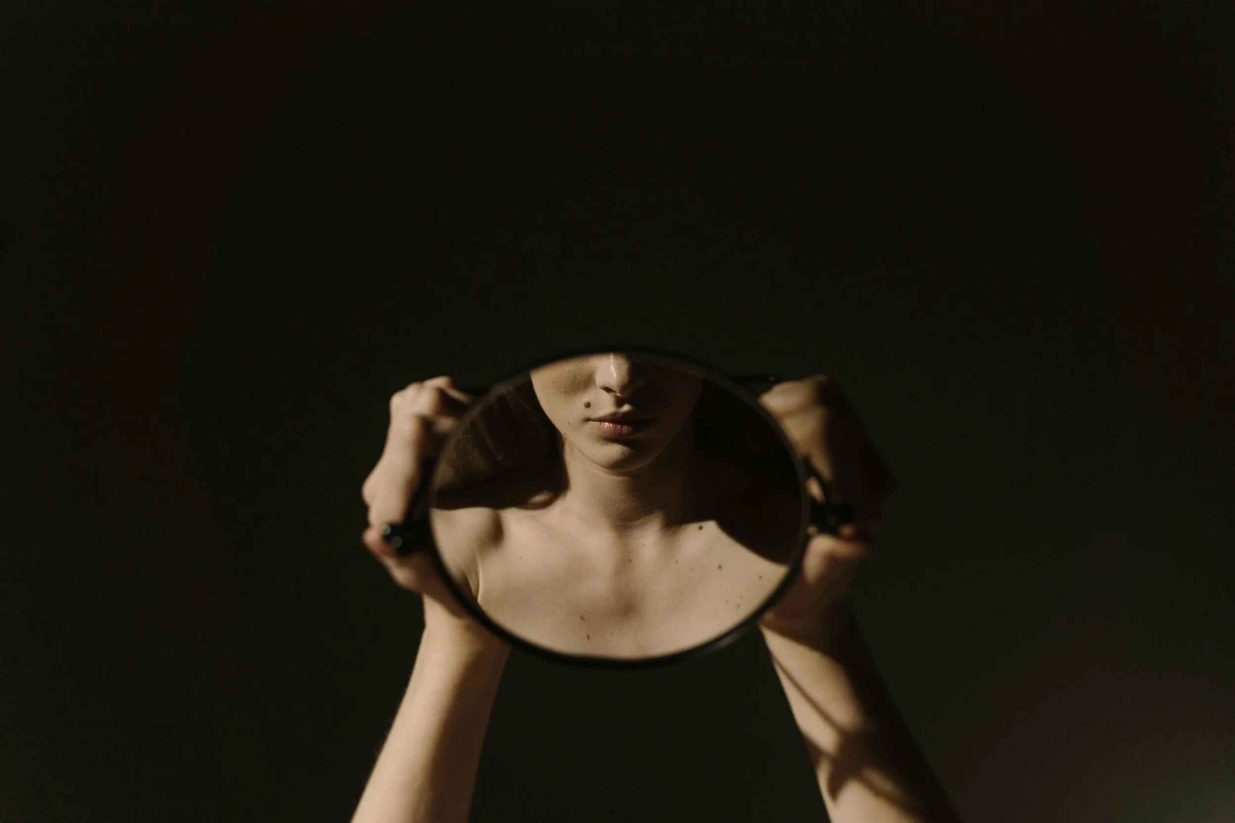 A woman holding a mirror in low light, symbolizing self-monitoring, identity, and the feminine wound of performance