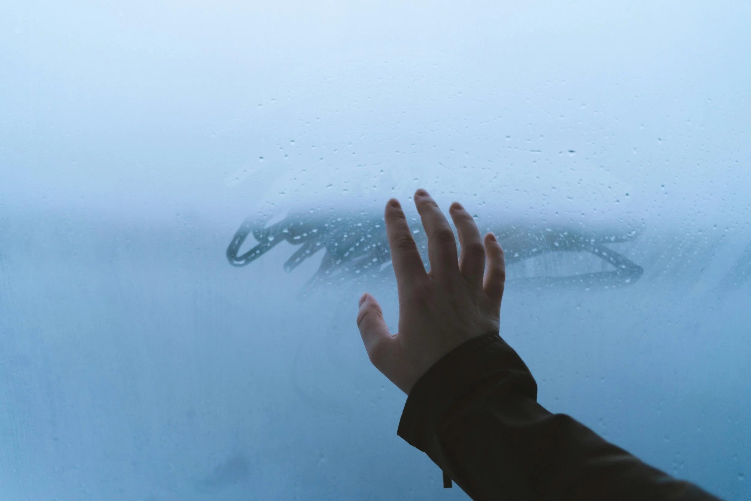 A hand resting on a fogged winter window, condensation softening the view outside, symbolizing breath, grounding, and nervous system settling.