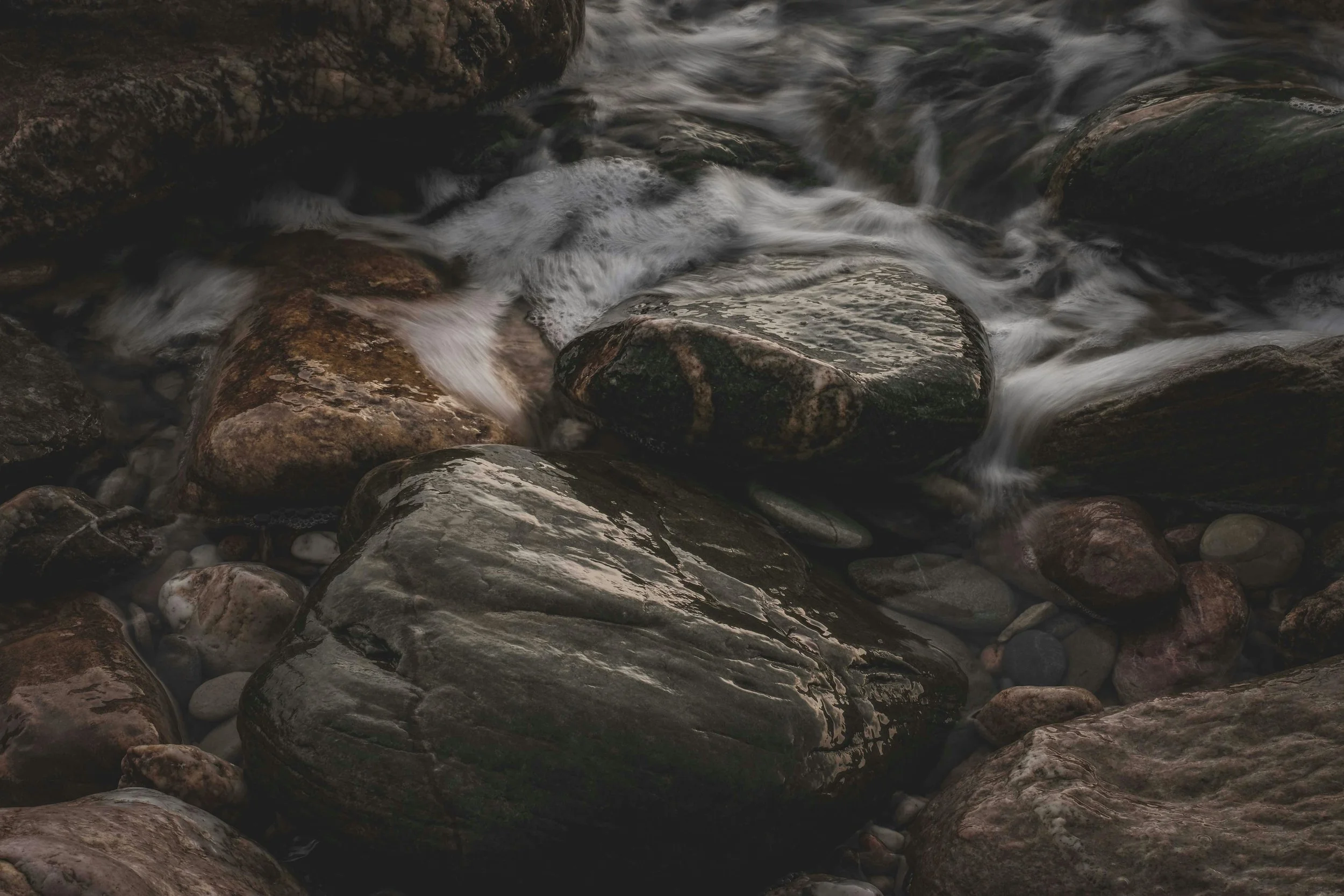 Dark, wet river stones in a rushing stream, capturing the slow, persistent force of water carving through rock.