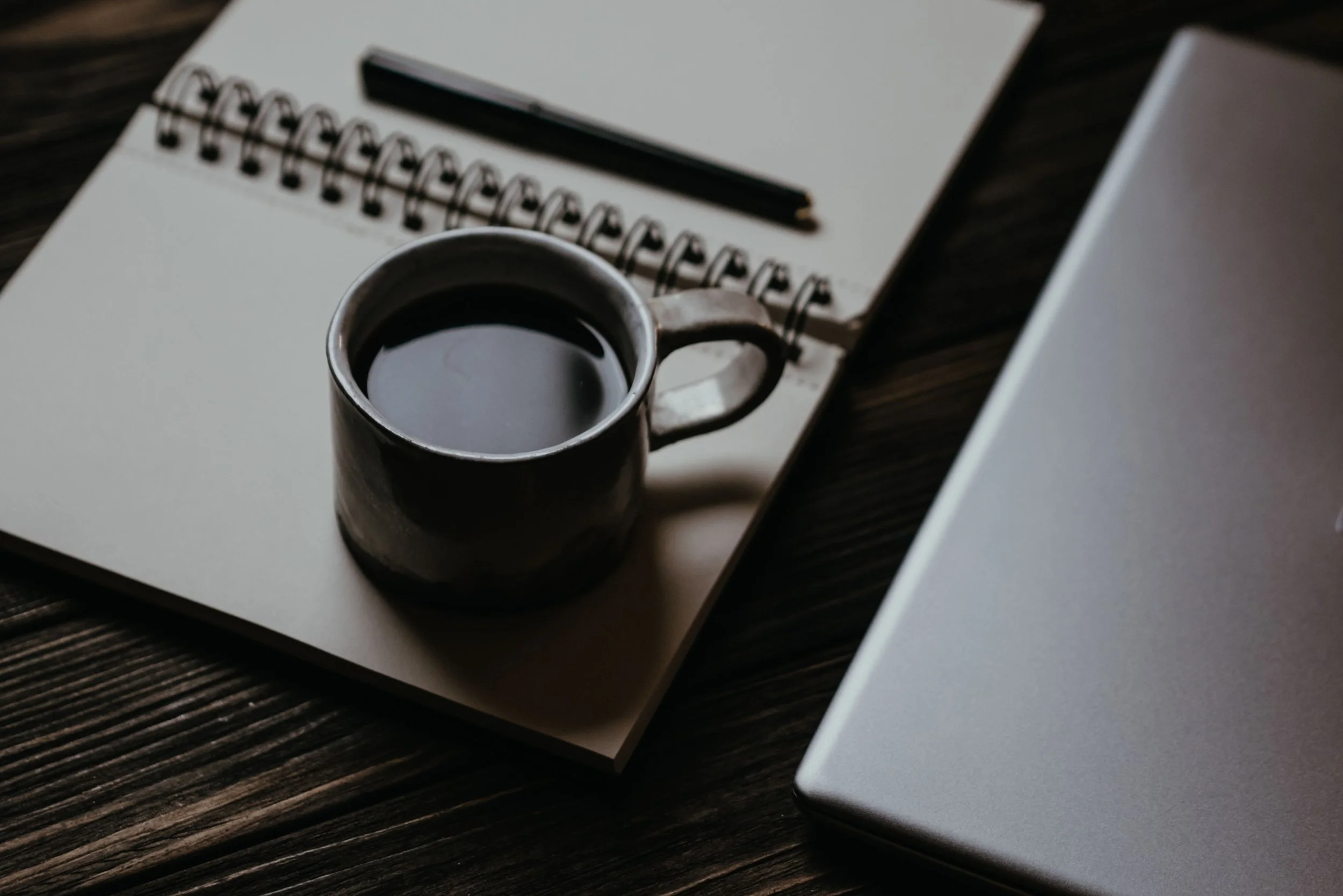 A moody, high-contrast photograph of a dark ceramic mug filled with liquid sitting on top of a spiral notebook, placed next to a sleek closed laptop on a dark wooden desk, capturing a quiet moment of dusk reflection or integration ritual.