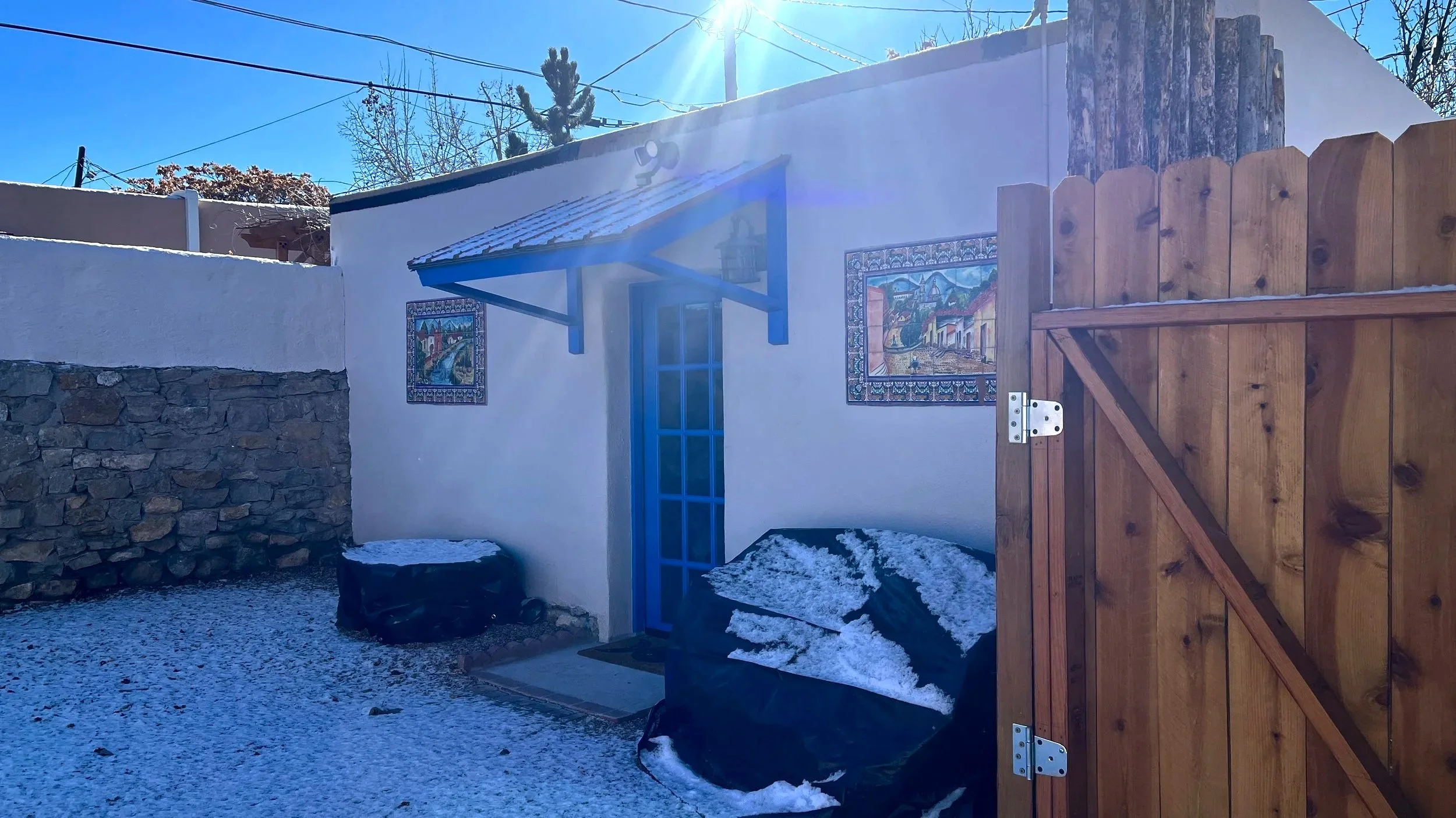 A quiet winter courtyard with a blue door, snow-covered ground, adobe walls, and soft morning light, creating a still and contemplative atmosphere.