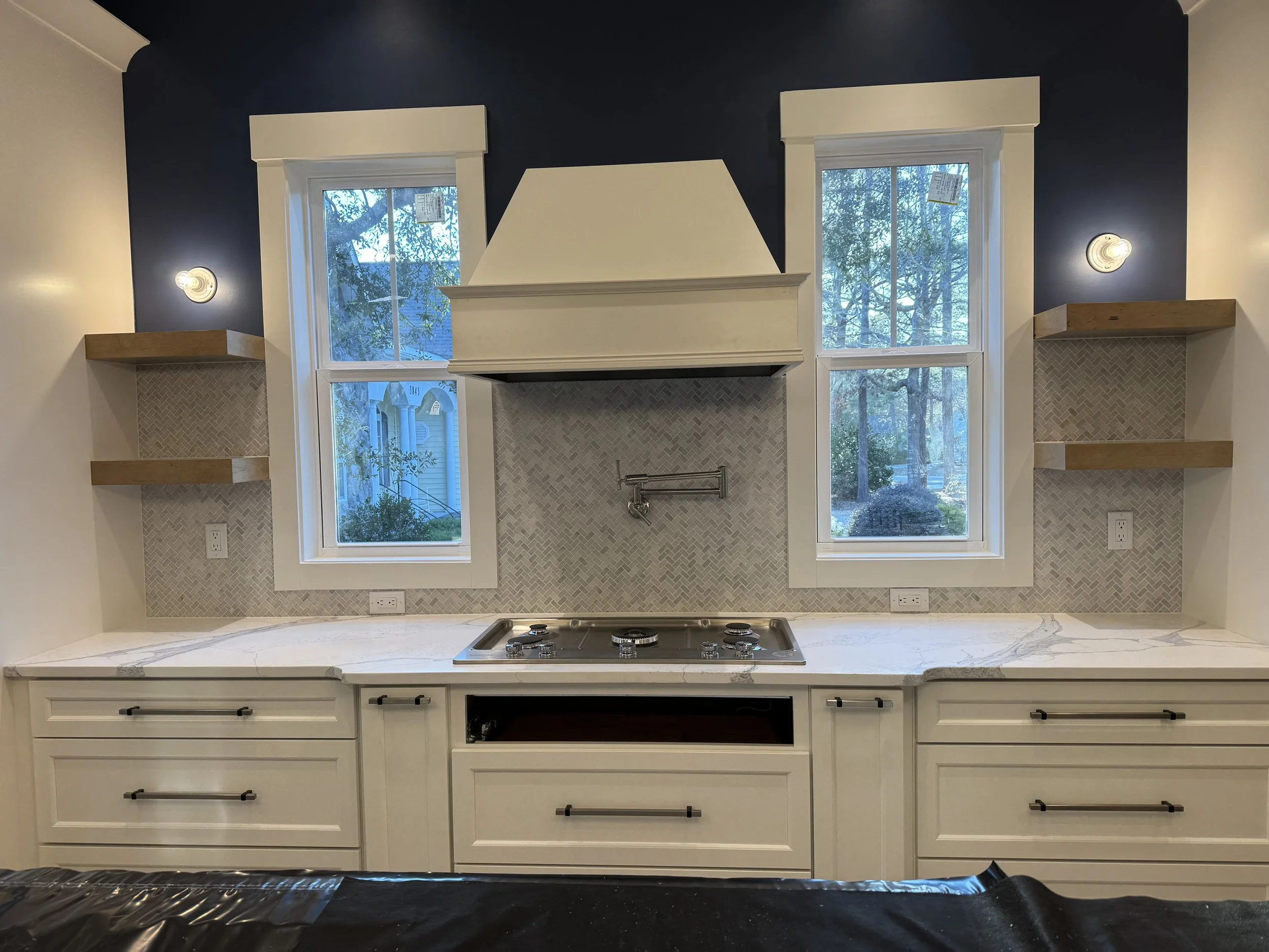A kitchen with white cabinets, a marble countertop, a stove, and two large windows with views of trees outside. There are built-in shelves on either side of the windows, wall-mounted lights, and a mosaic tile backsplash.