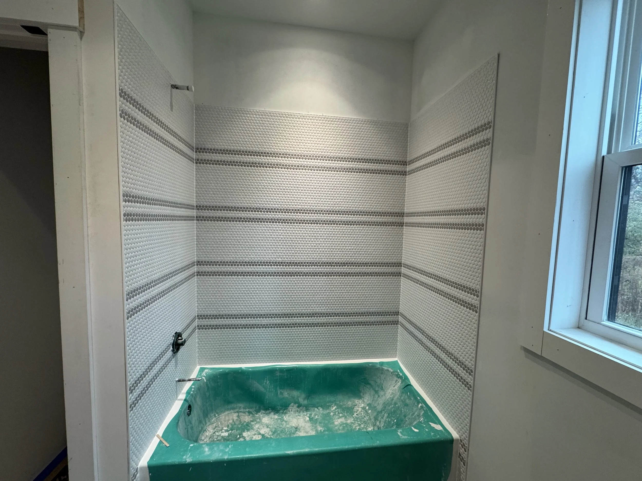 Bathroom under construction with partially tiled wall and a green bathtub covered in protective plastic.